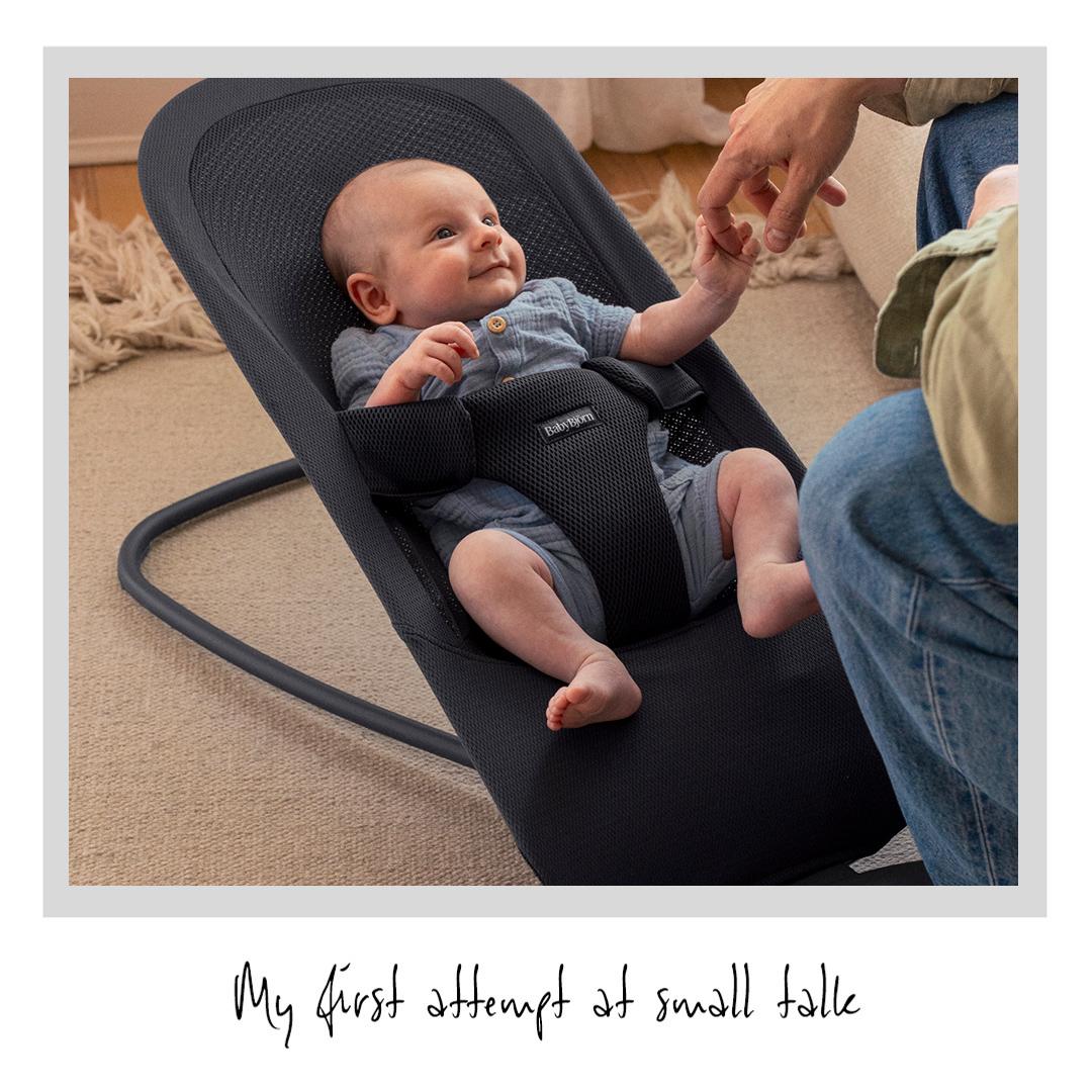A baby in a bouncer seat reaches for an adult's hand. A handwritten caption below reads, "My first attempt at small talk."