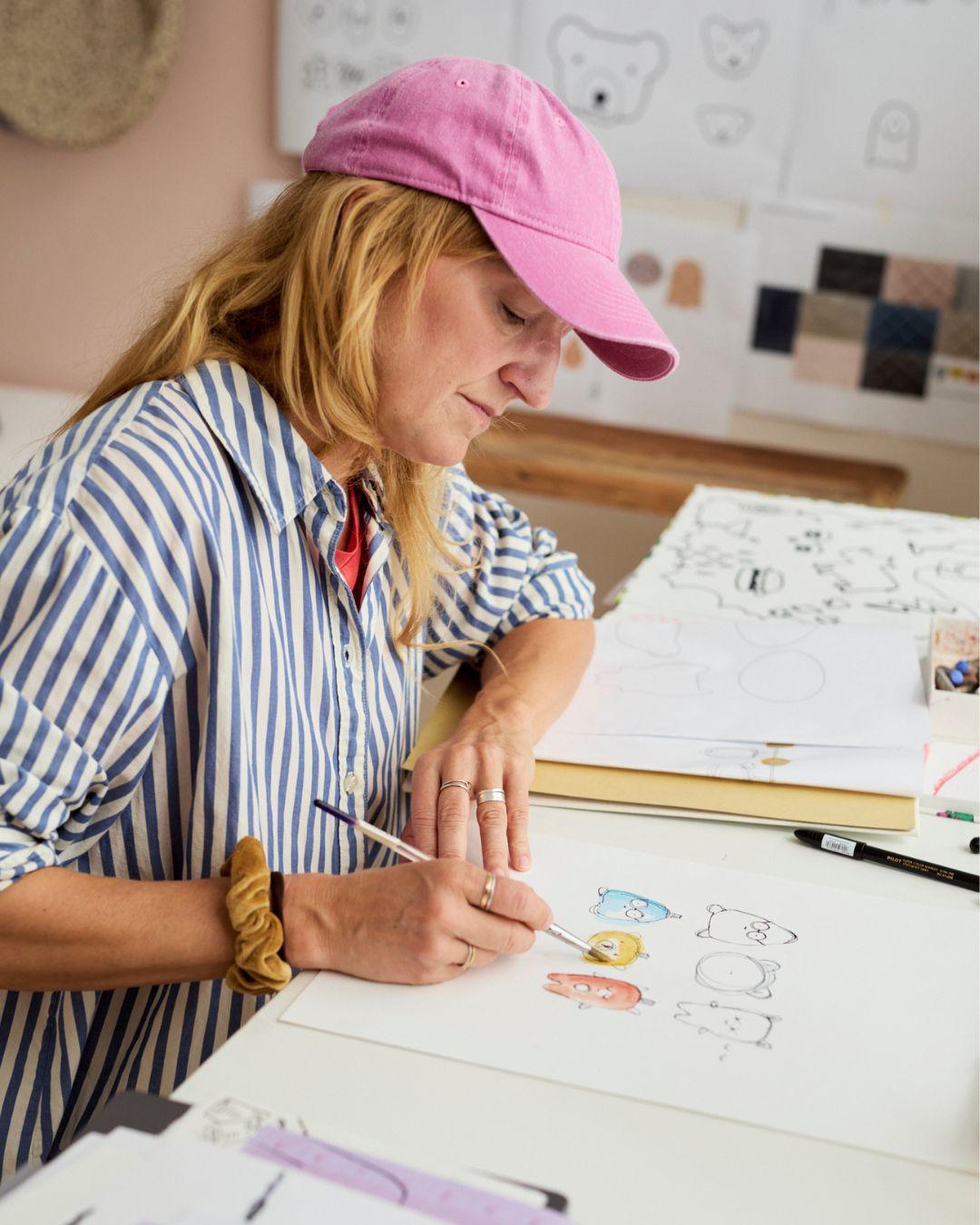 Person in a striped shirt and pink cap drawing cartoon animals at a desk with sketches on the wall.
