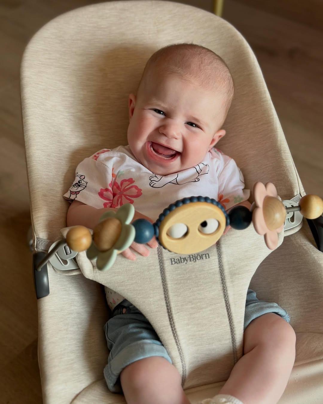 Smiling baby sitting in a beige BabyBjörn bouncer, wearing a floral shirt and denim shorts, with a toy bar in front.