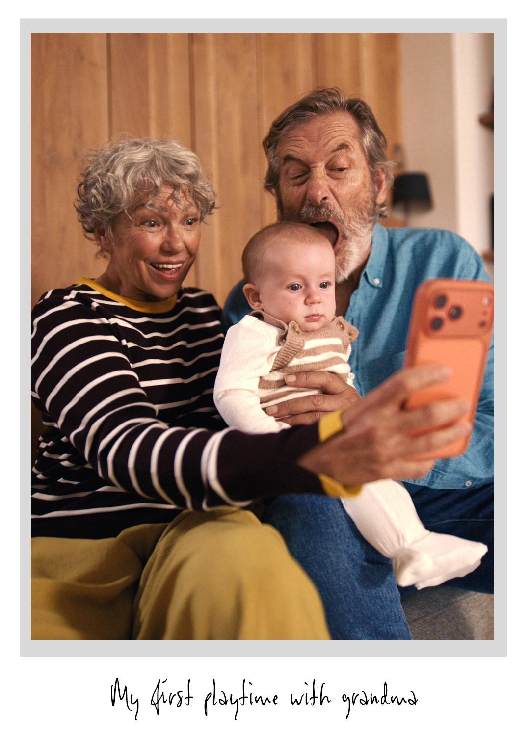 Smiling elderly couple and a baby taking a selfie with a smartphone on a couch. The photo caption reads: "My first playtime with grandma."