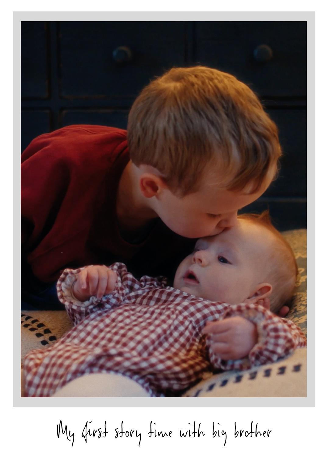A young boy gently kisses a baby lying down, both wearing red checkered outfits. The photo caption reads, "My first story time with big brother."