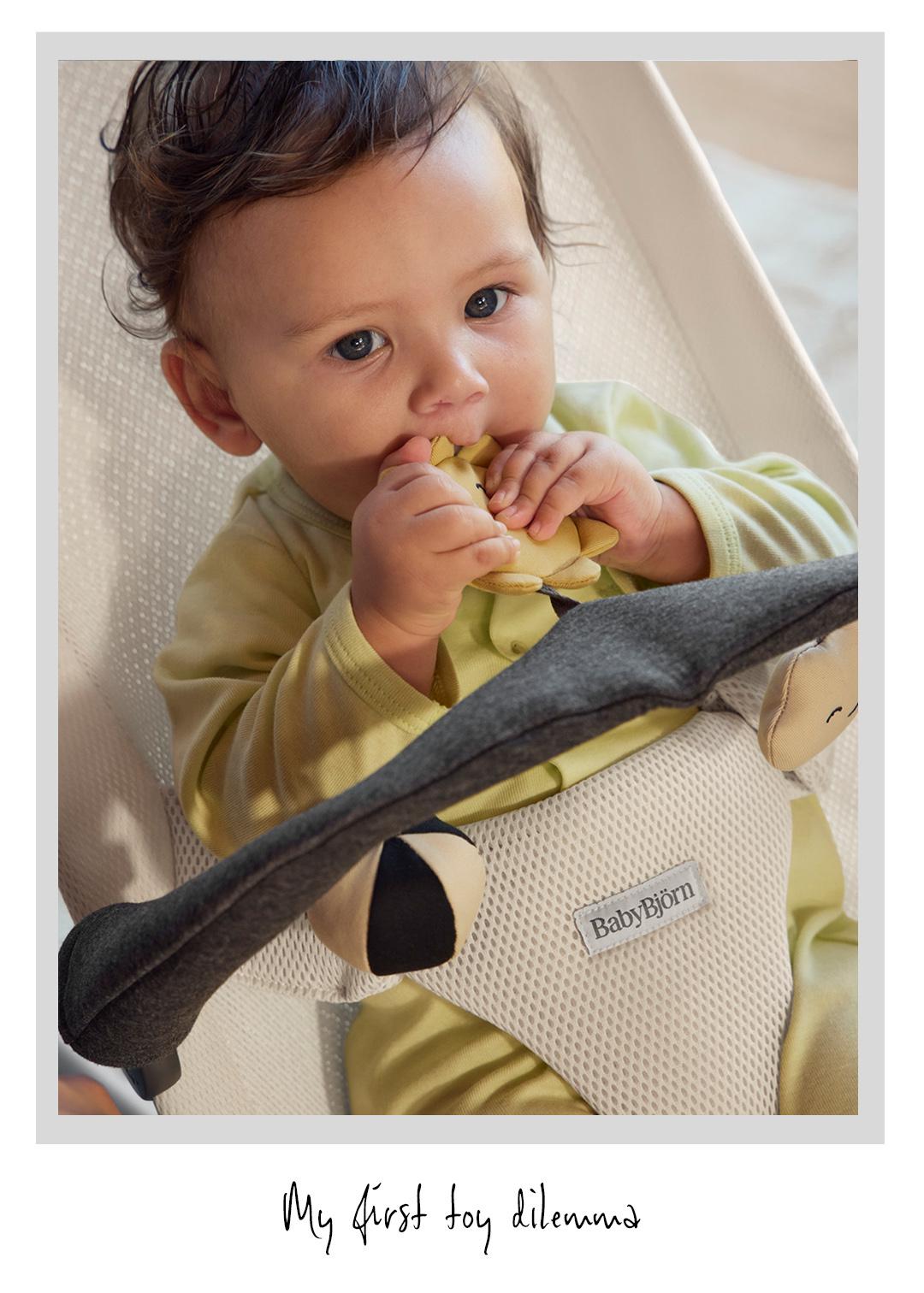 A baby in a yellow outfit sits in a BabyBjörn bouncer, holding a toy and looking up with curiosity. Text below reads "My first toy dilemma."