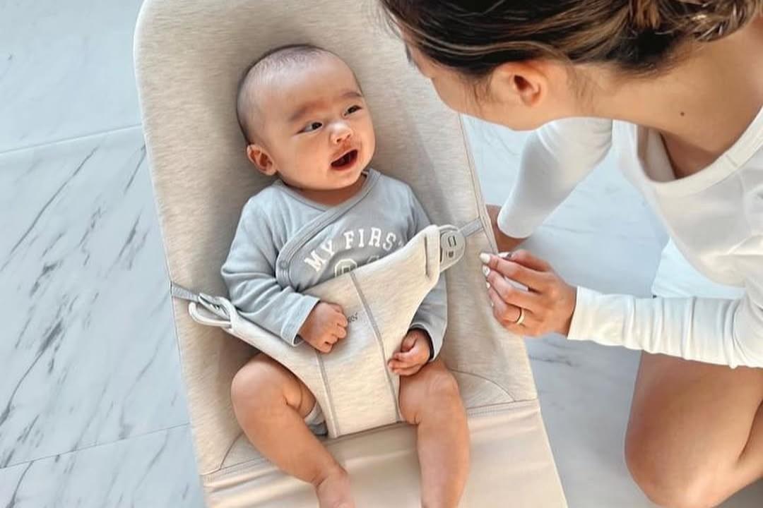A baby smiling while seated in a beige baby chair, wearing a gray outfit, looking up at an adult kneeling nearby on a light-colored floor.