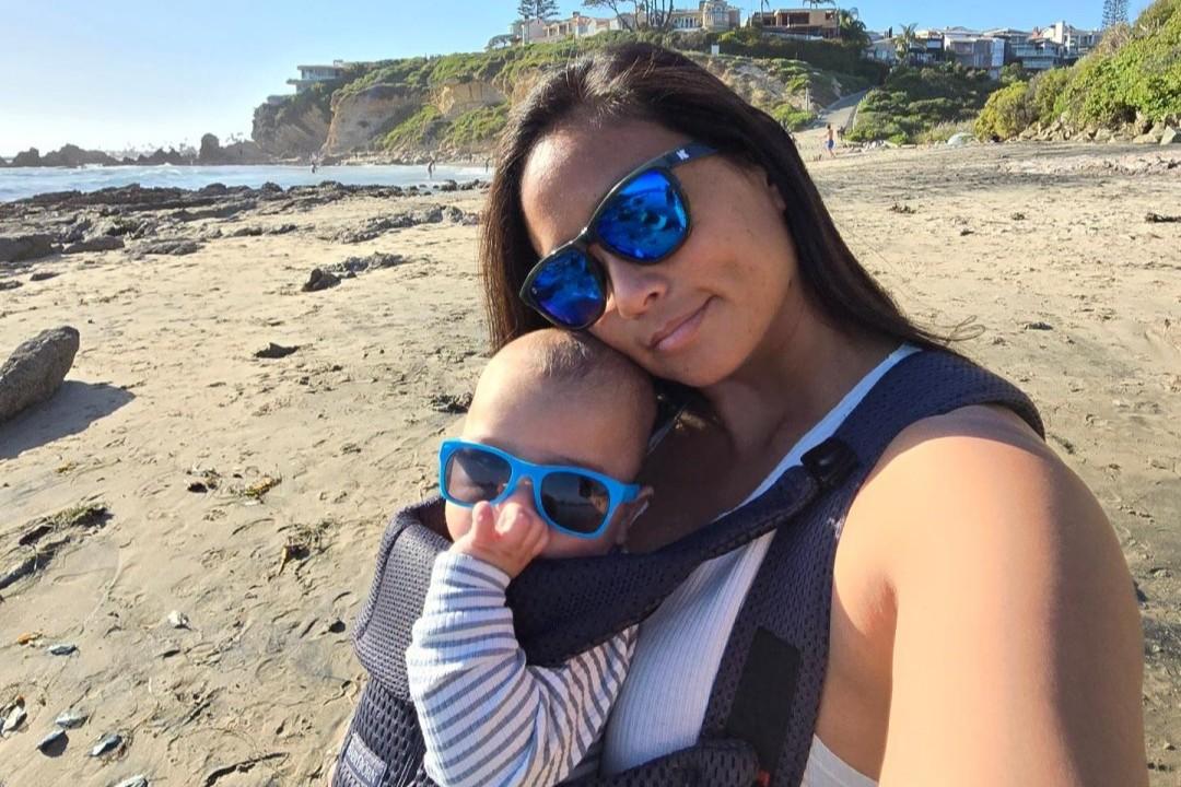 A woman wearing sunglasses takes a selfie on a sandy beach while carrying a baby in a carrier. The baby is also wearing sunglasses.