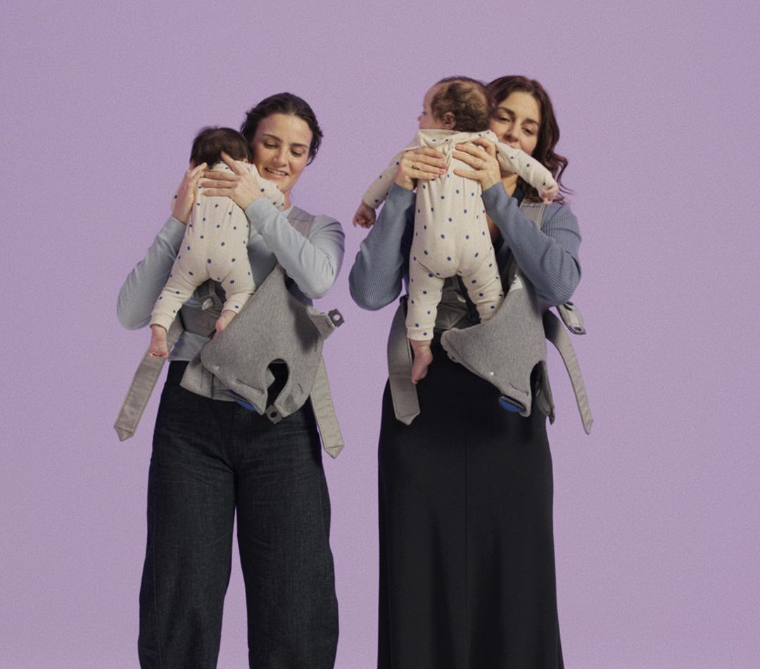 Two women hold babies with star-patterned onesies, using grey baby carriers against a lavender background.