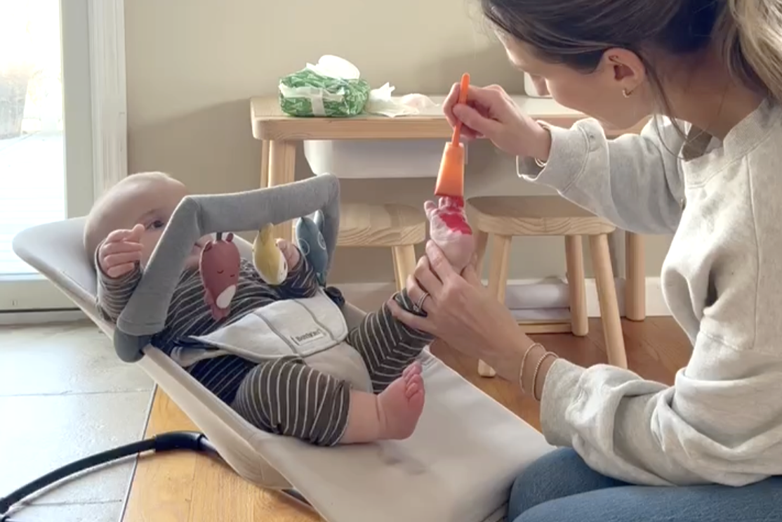 A baby sitting in a bouncer is being fed with a spoon by a woman in a cozy room with a table in the background.