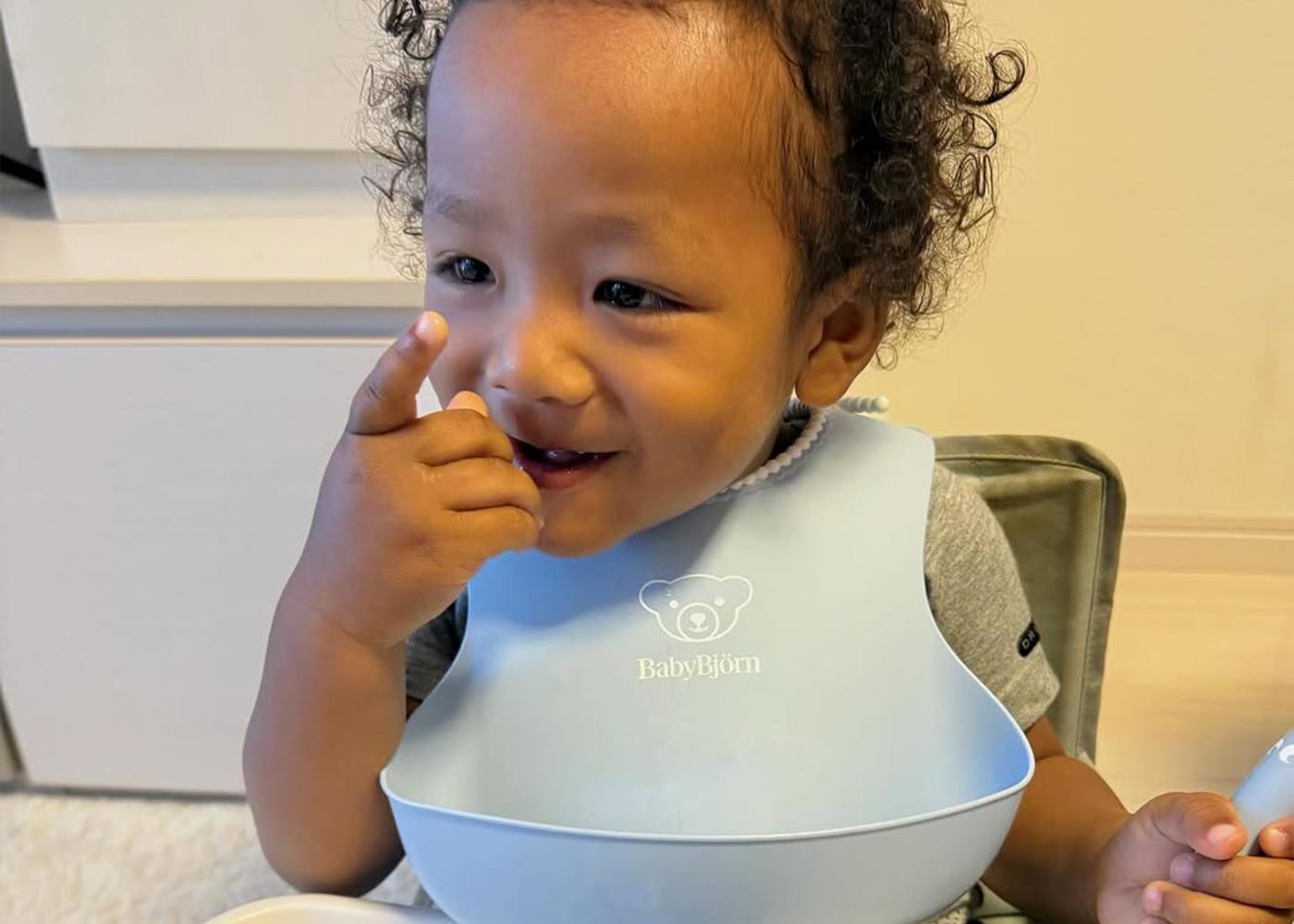 A toddler wearing a blue bib sits at a table, smiling and pointing with one hand. A bowl is in front of them.