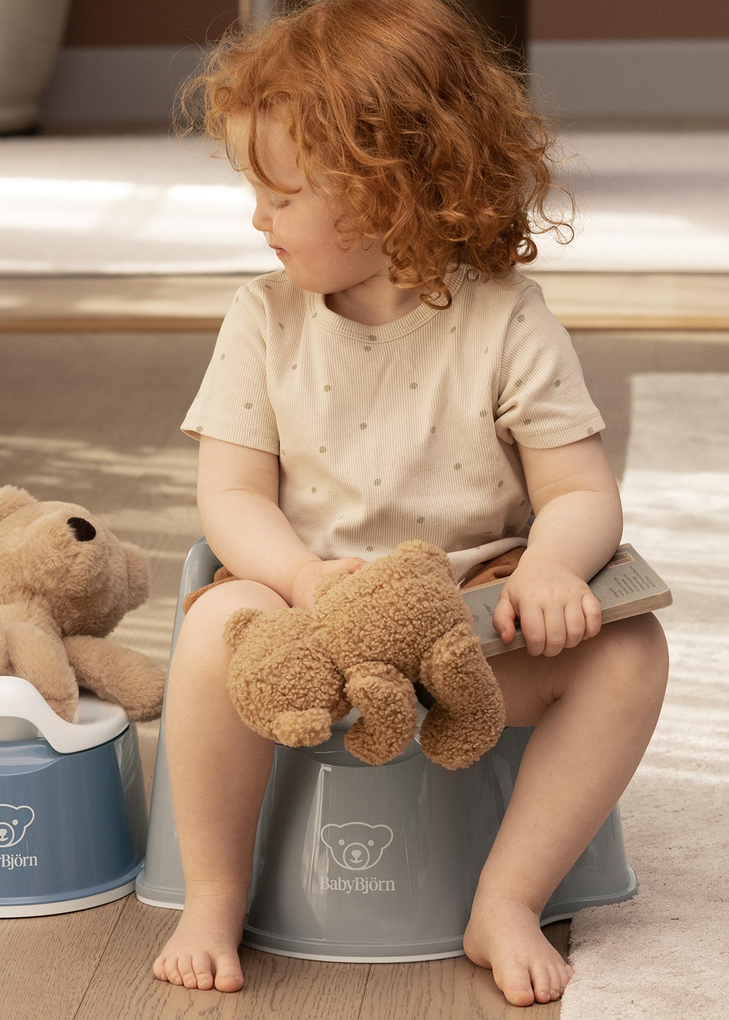A toddler with curly red hair sits on a blue potty, holding a teddy bear and a small book, looking to the side.