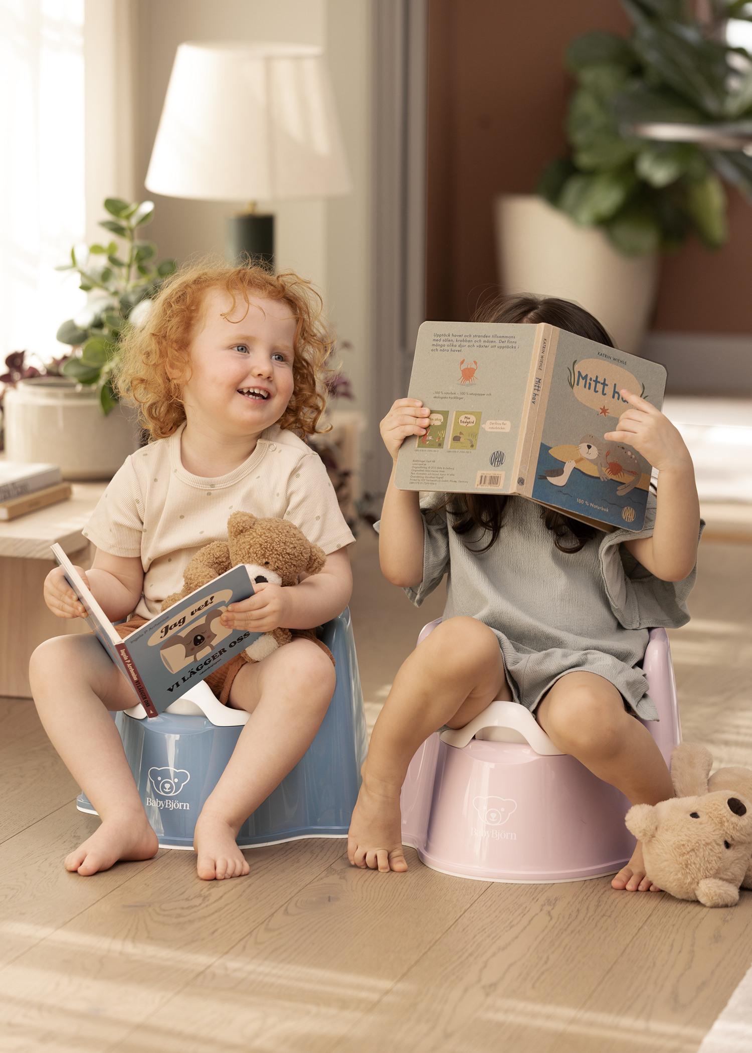Two children sitting on potties, one reading a book and the other holding a teddy bear, in a cozy room with plants and soft lighting.