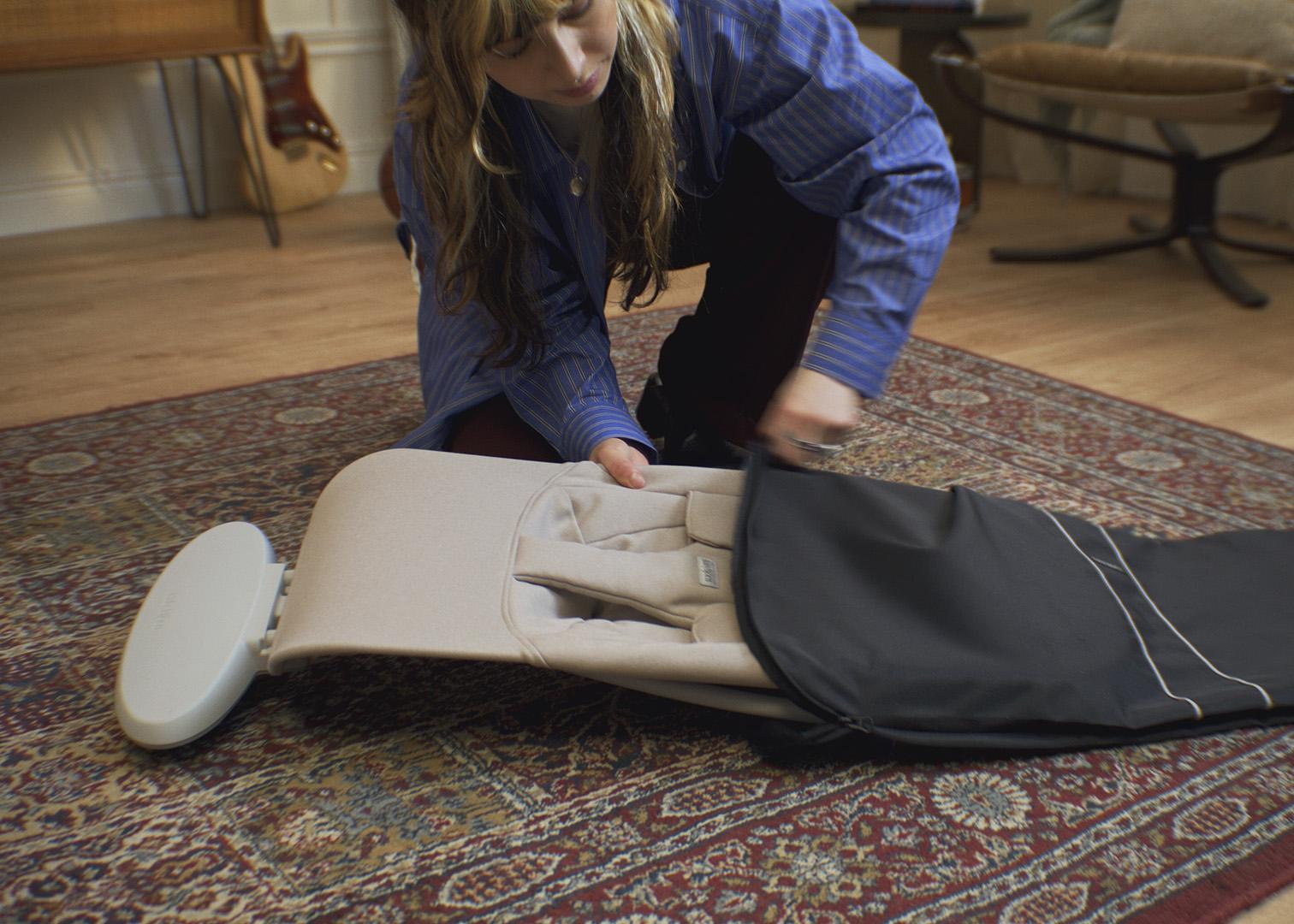 A person folding a baby bouncer on a patterned rug in a cozy living room setting.