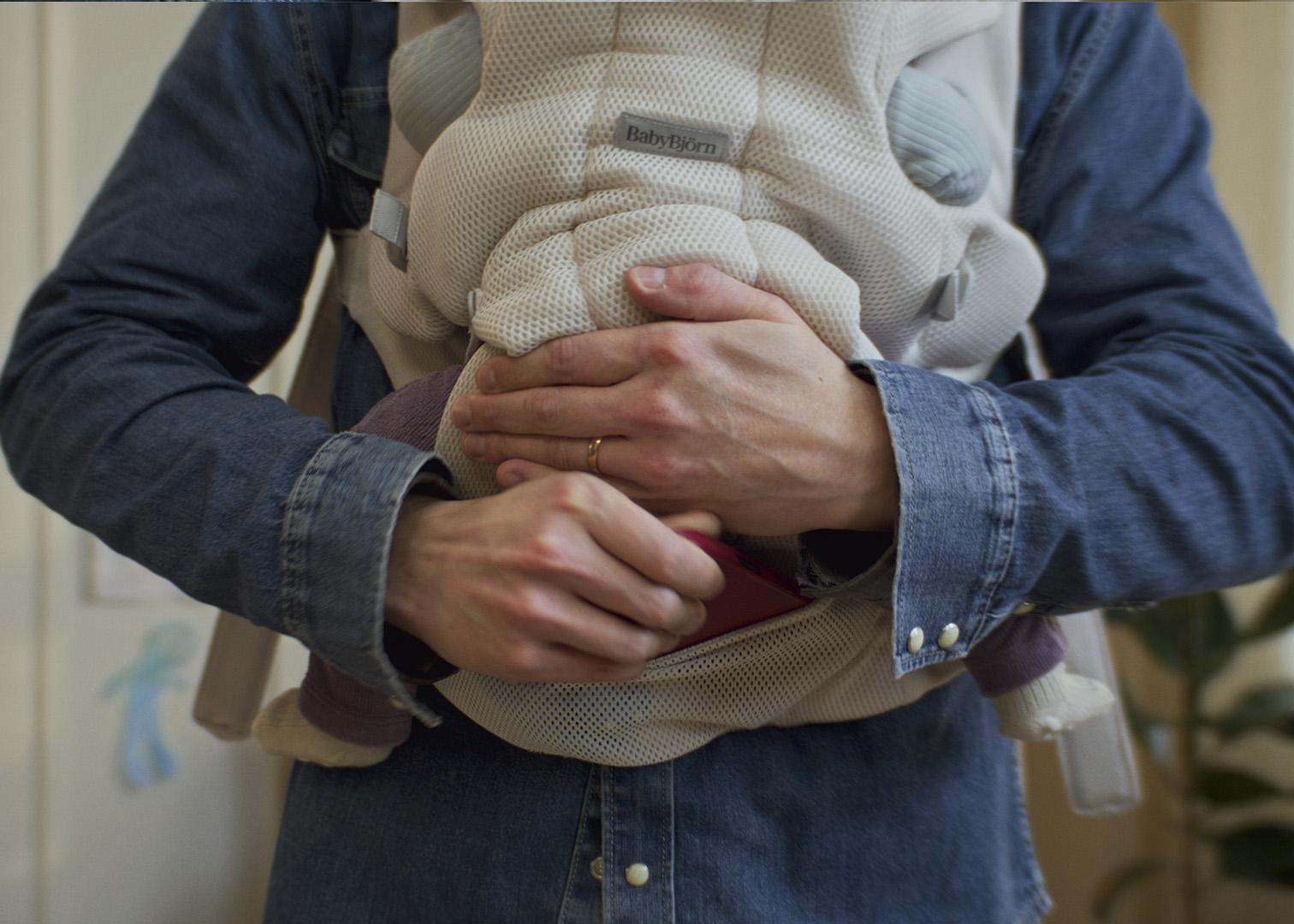 Person wearing a denim shirt holds a baby in a front-facing carrier, hands gently supporting the child, indoors.