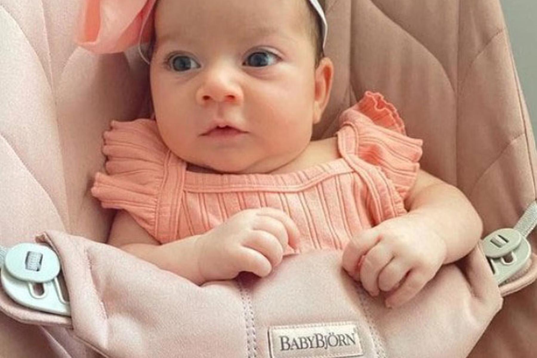 Baby in a pink dress and headband sitting in a BabyBjörn seat, looking alert and curious.