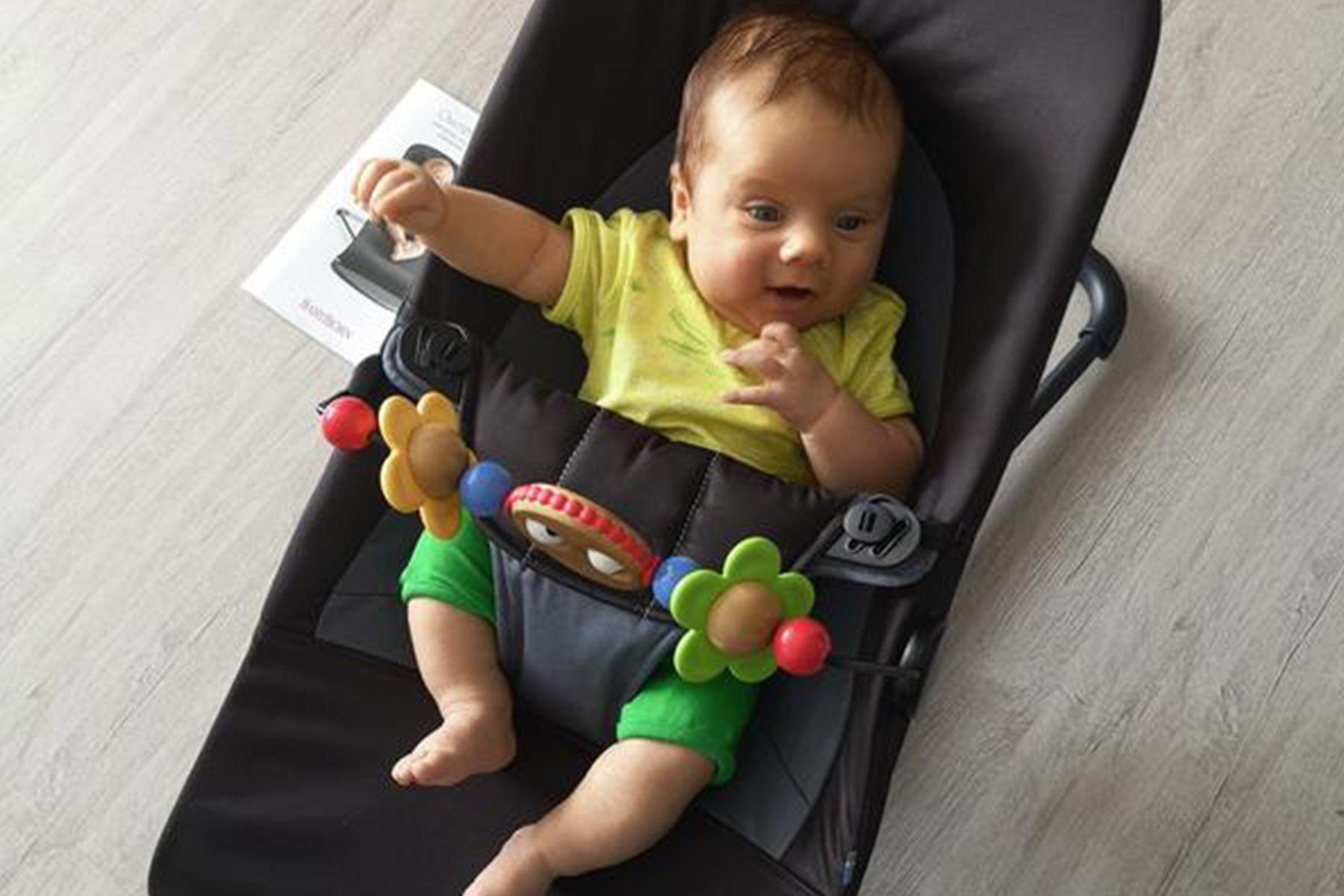 Infant in a black bouncer, wearing a yellow shirt and green pants, reaching for a colorful toy bar with flower shapes.