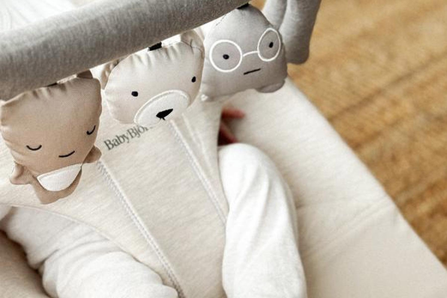 Infant in white sleeper beneath a toy bar with three hanging plush animal faces: sleeping, bear, and bespectacled gray.