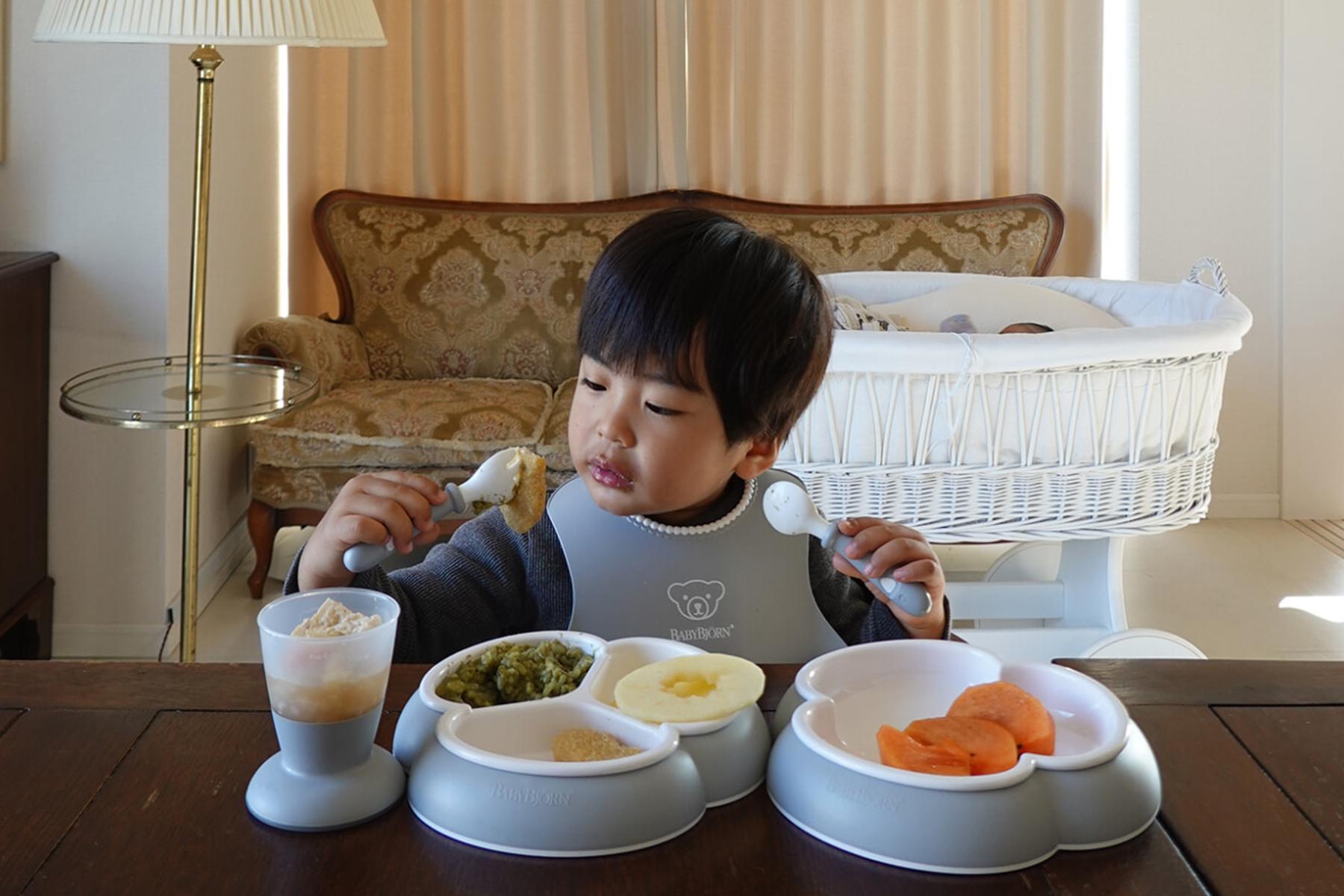 A child in a high chair eats with a spoon, surrounded by plates of pureed food, in a cozy living room setting.