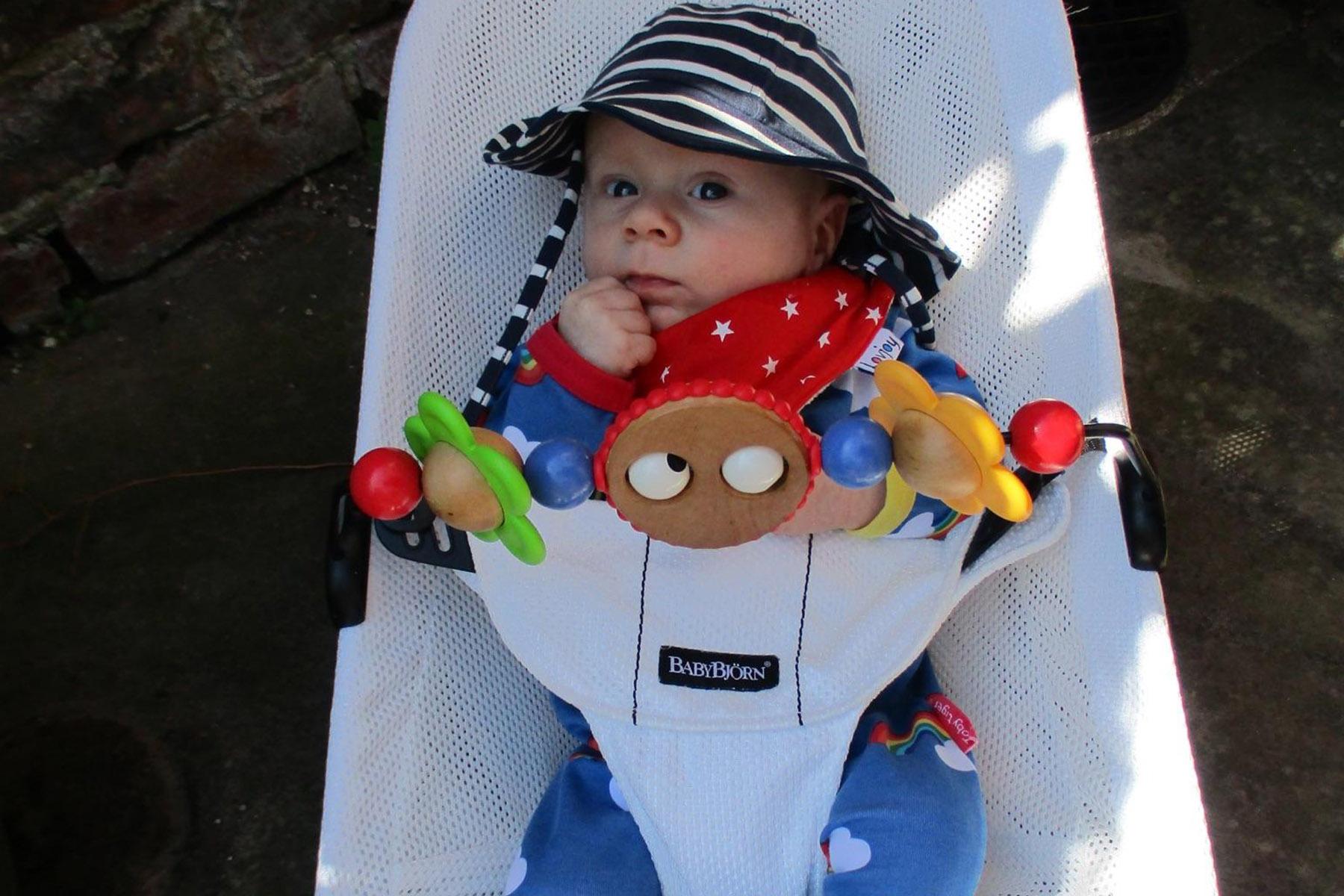 Baby in white bouncer wearing striped sunhat and red star bib, chewing finger and holding colorful toy bar.