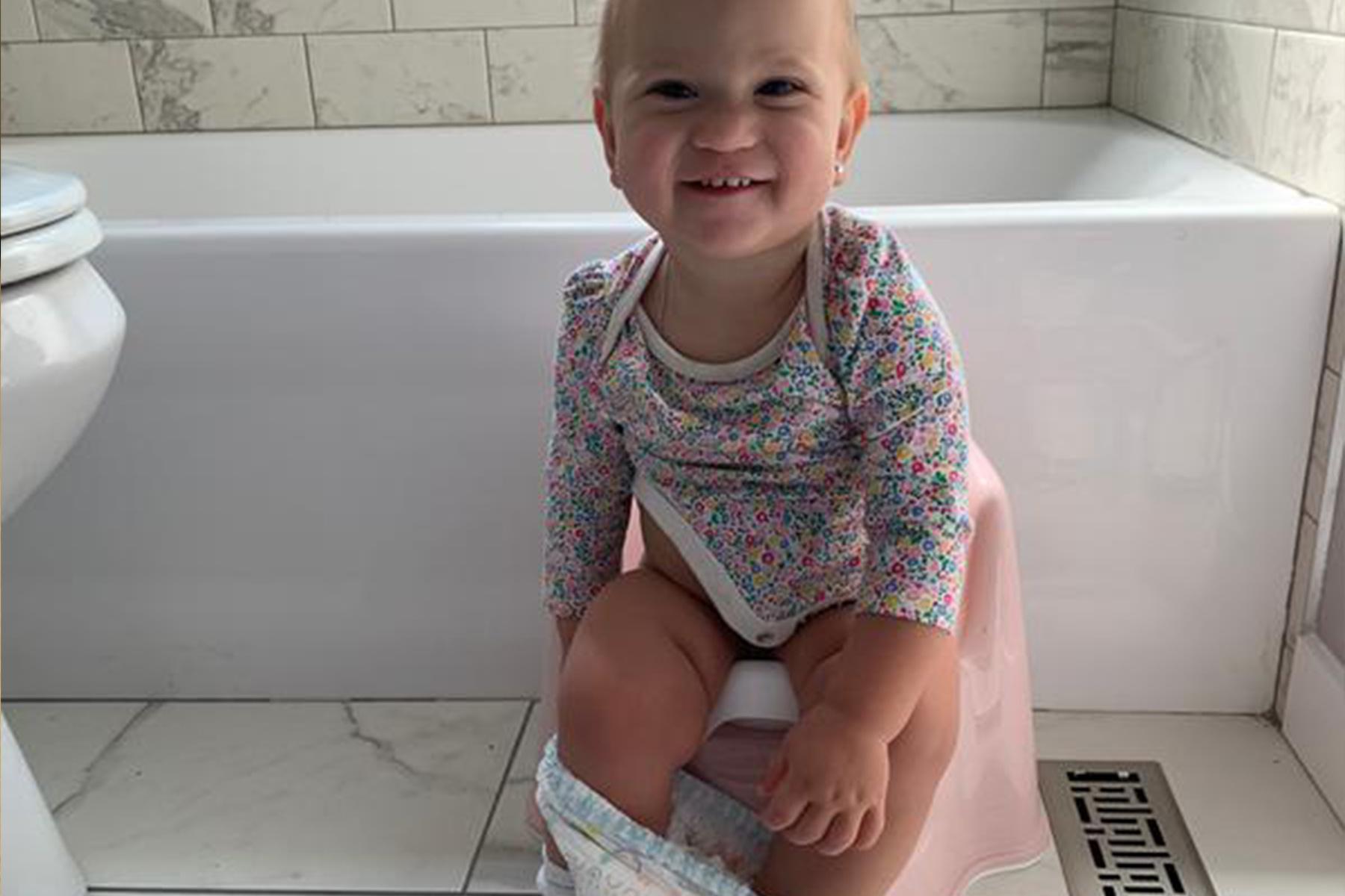 Smiling toddler in a floral onesie sitting on a pink potty in a tiled bathroom, underwear pulled down around the ankles.