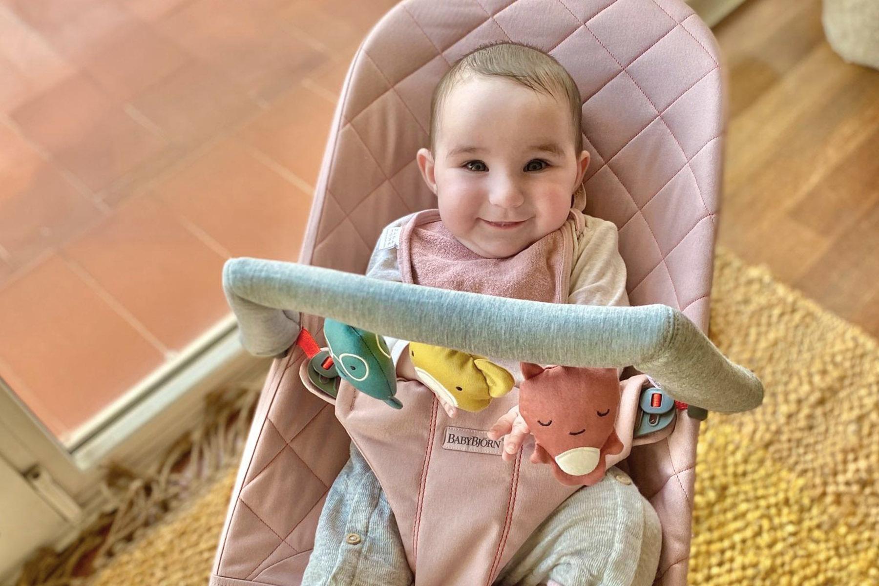 Smiling baby in a pink quilted bouncer wearing a bib, with a toy bar of plush animals across the front.