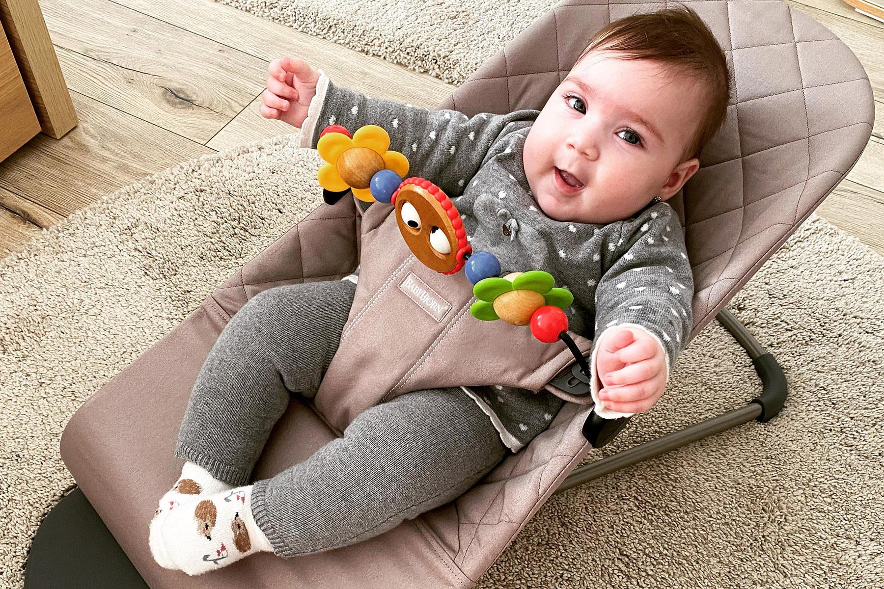 Smiling baby in gray outfit reclining in a beige bouncer, holding a colorful toy bar with flowers and beads.