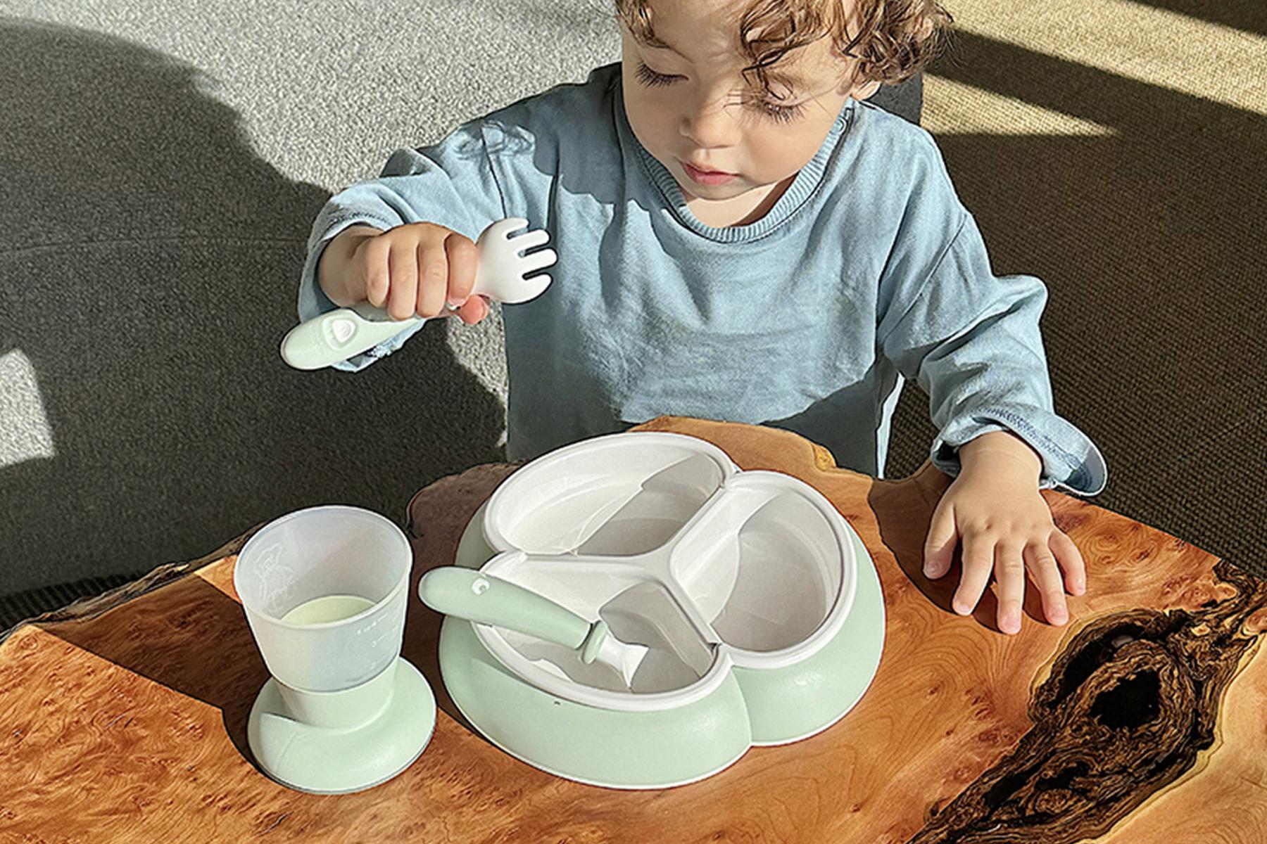 A toddler in a blue shirt uses a fork with a divided plate on a wooden table, next to a cup.