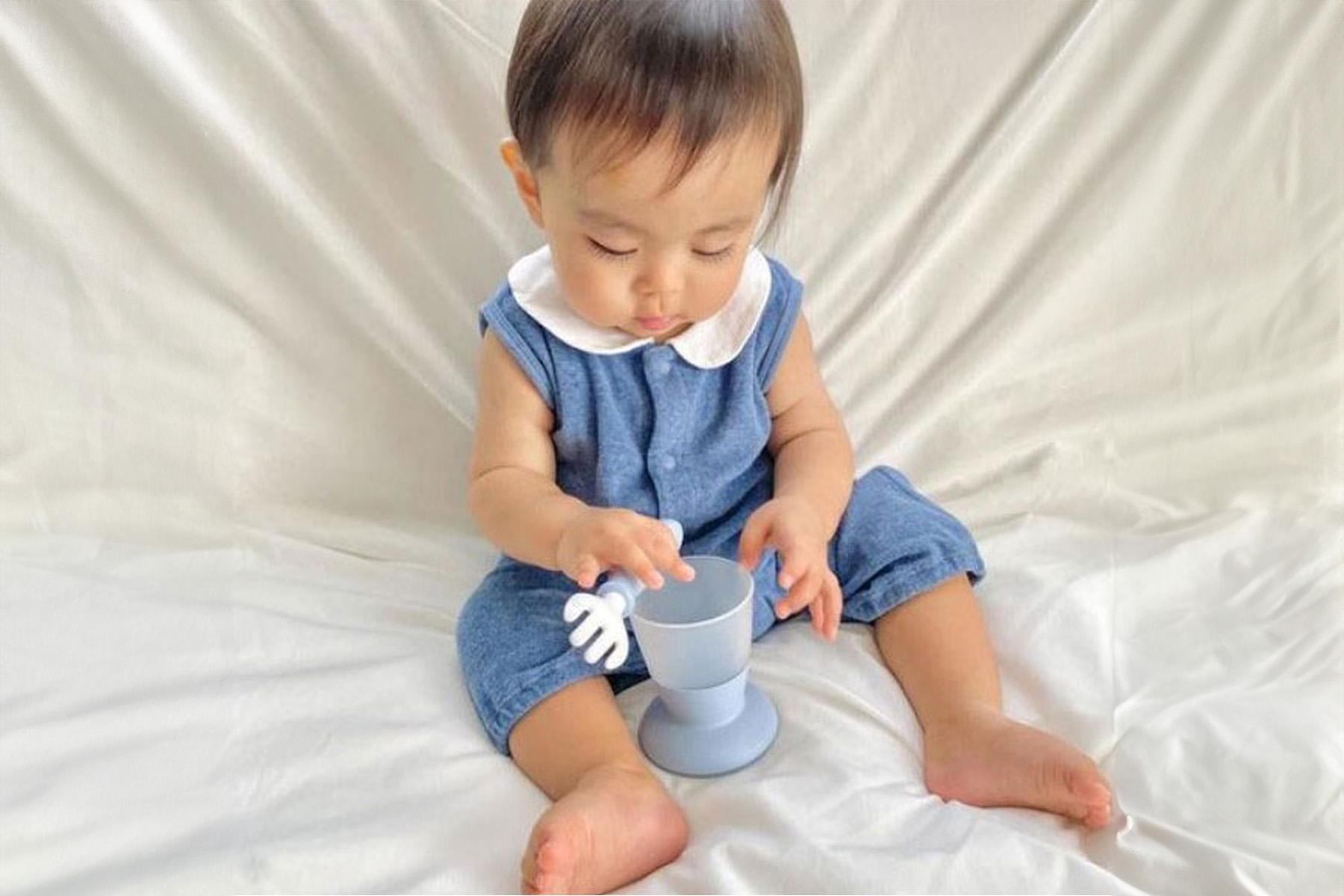 A baby in a denim outfit sits on a white blanket, playing with a blue cup that has a white handle.