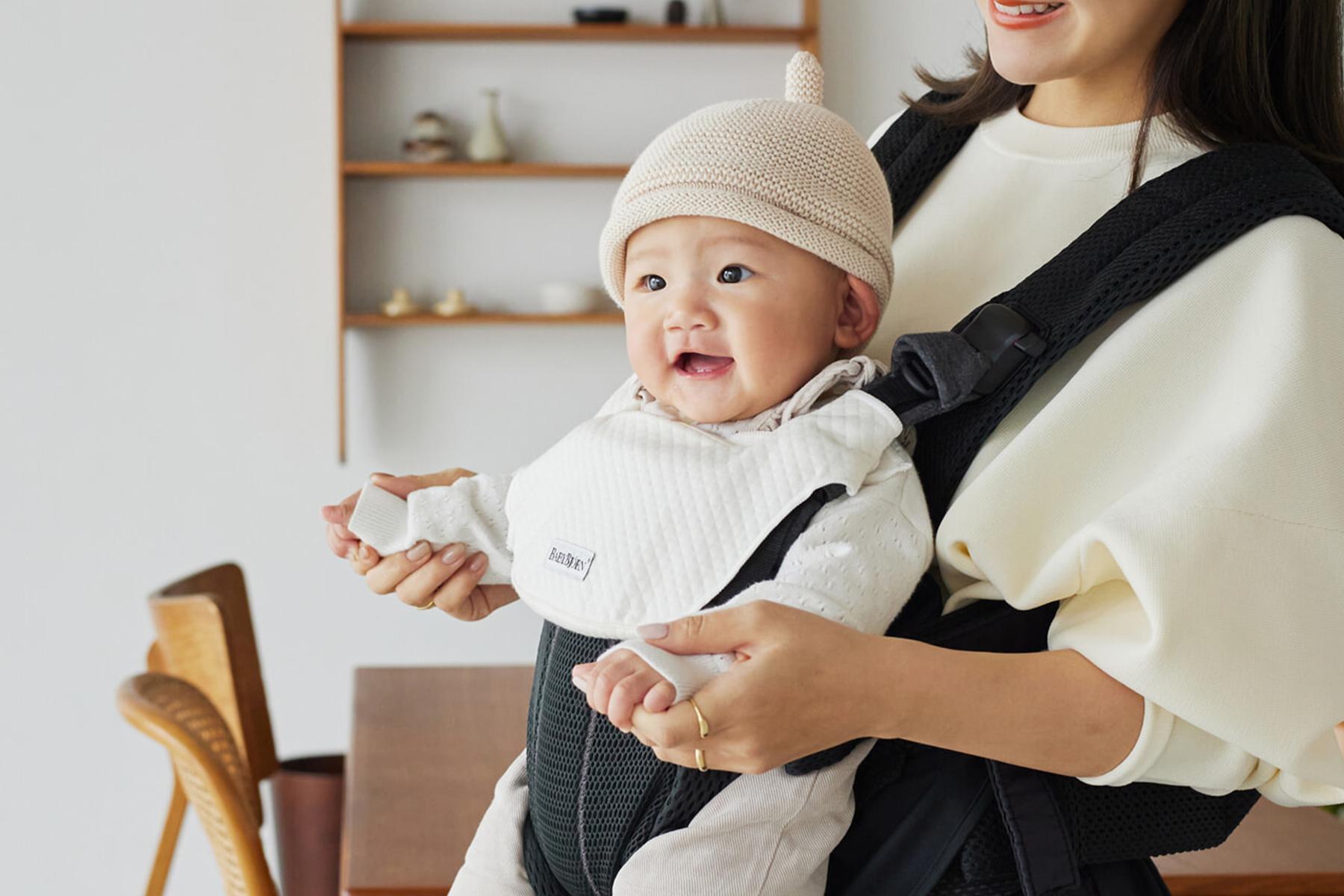 A smiling baby in a beige hat is held by a woman wearing a white sweater and black baby carrier, in a cozy room with wooden shelves.