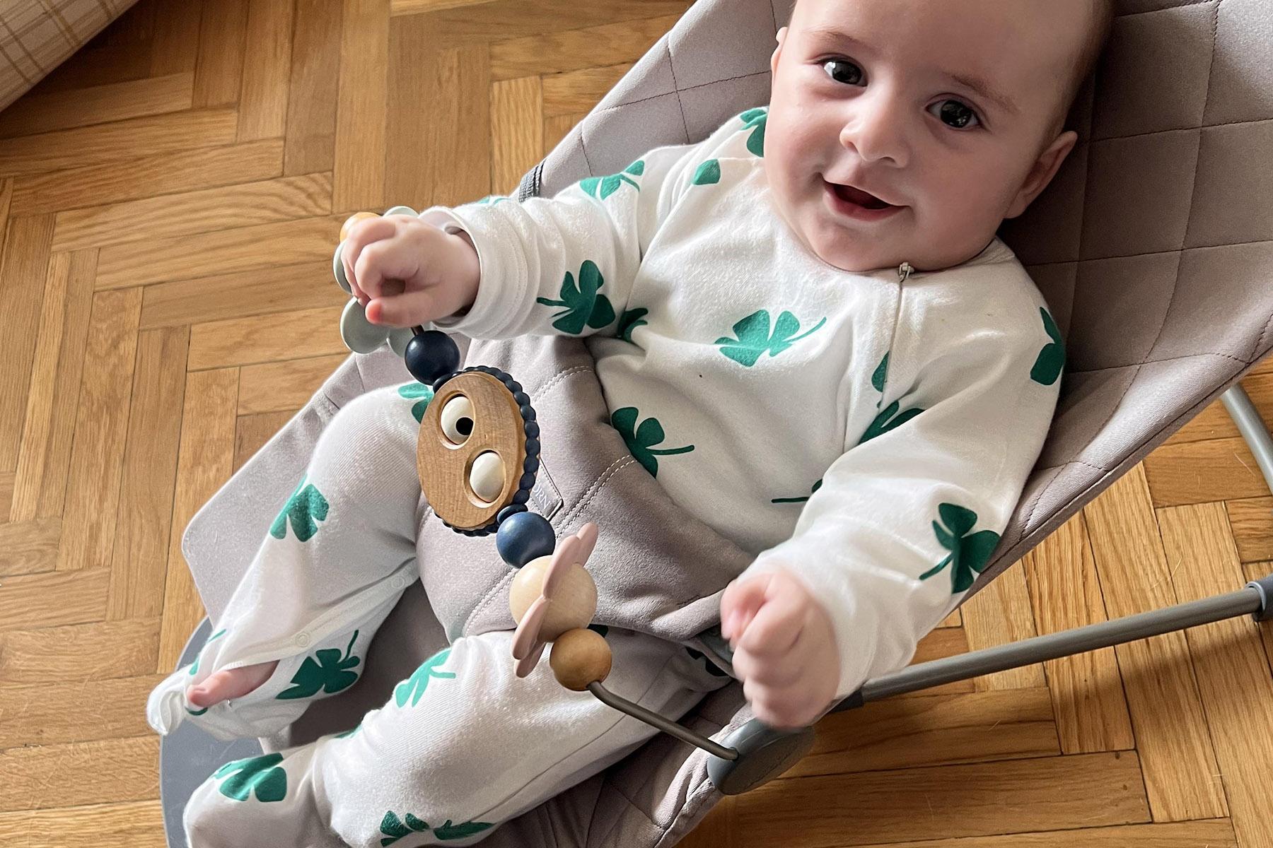 Smiling baby in white onesie with green clover prints sits in a gray bouncer holding a wooden lion toy on a parquet floor.