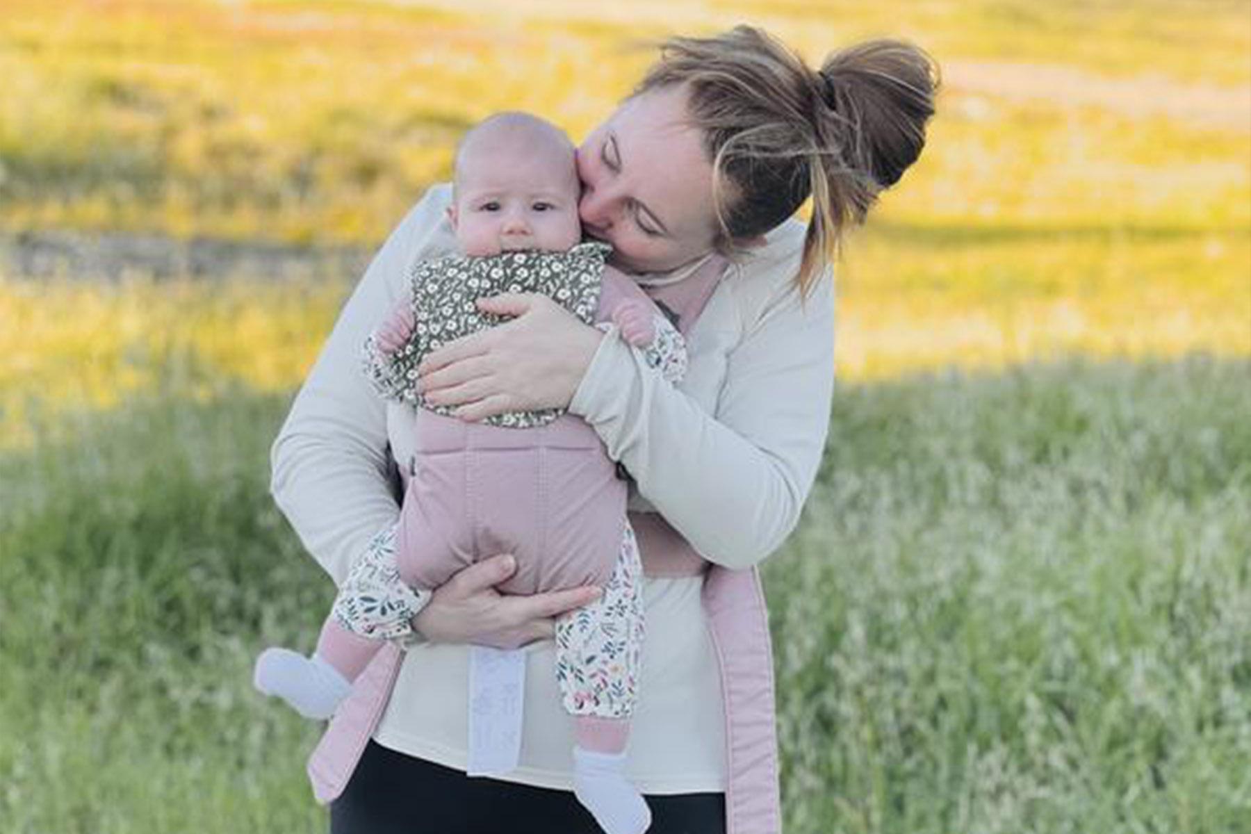 Woman cradling and kissing a baby in a grassy field; infant facing and looking toward the camera.