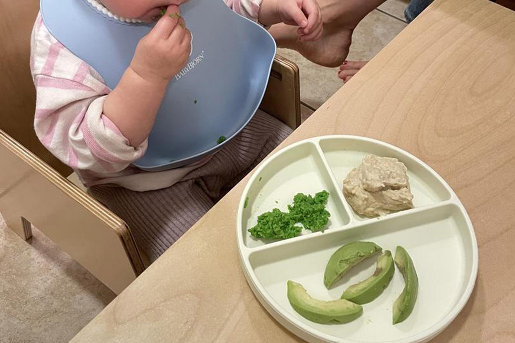 Baby in a high chair wearing a blue bib, picking at food; divided plate with avocado slices, mashed food and chopped greens.