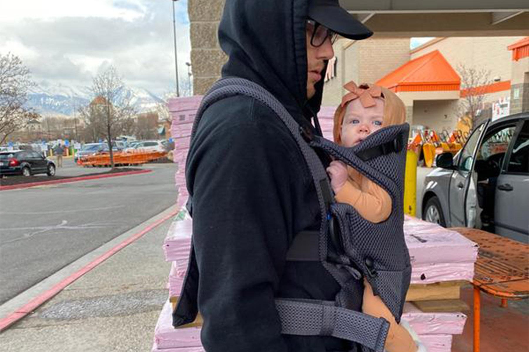 Adult in a black hoodie and cap carrying a baby with a pink bow in a front carrier outside a store.