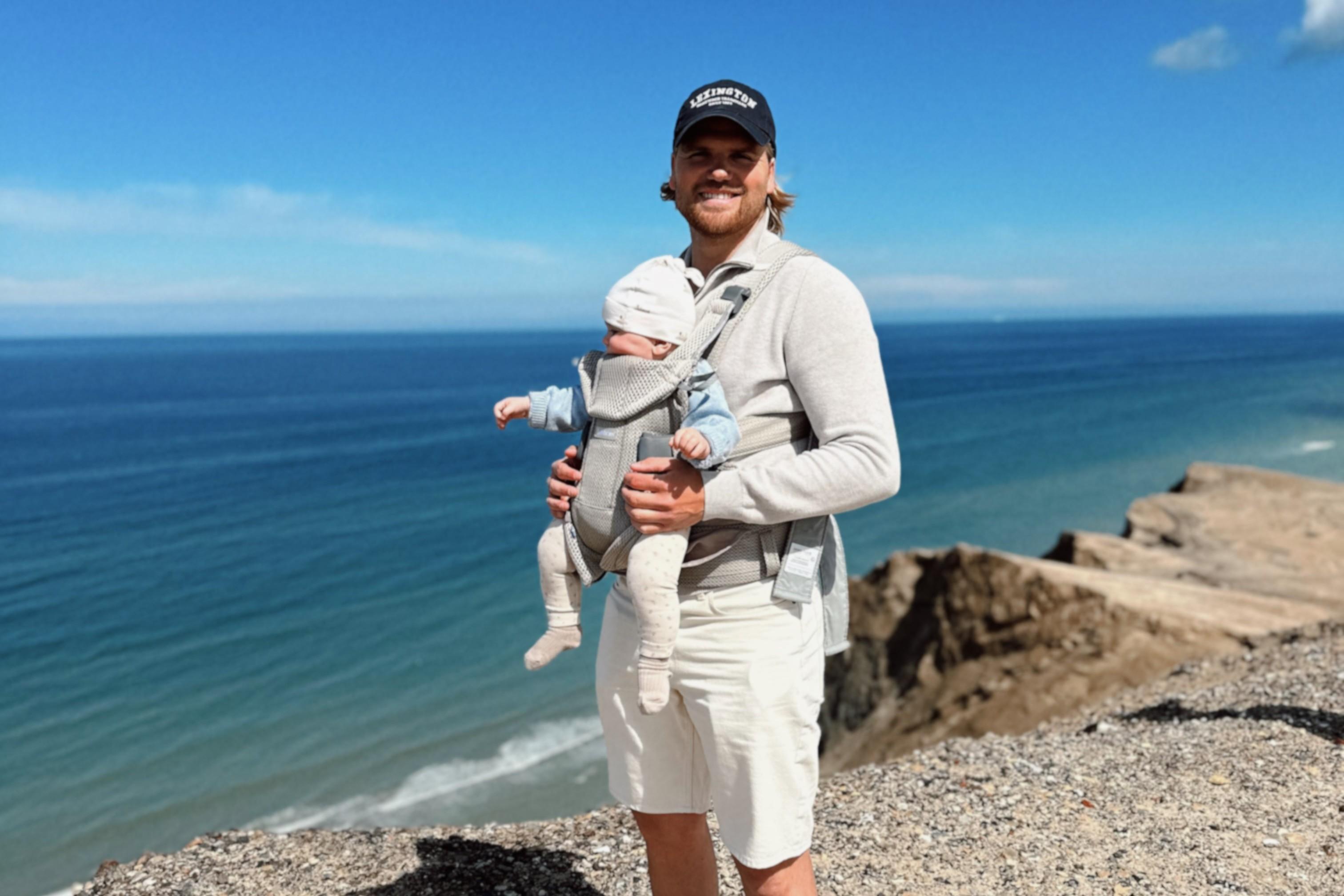 Man wearing a cap holds a baby in a carrier by a seaside cliff, under a clear blue sky.