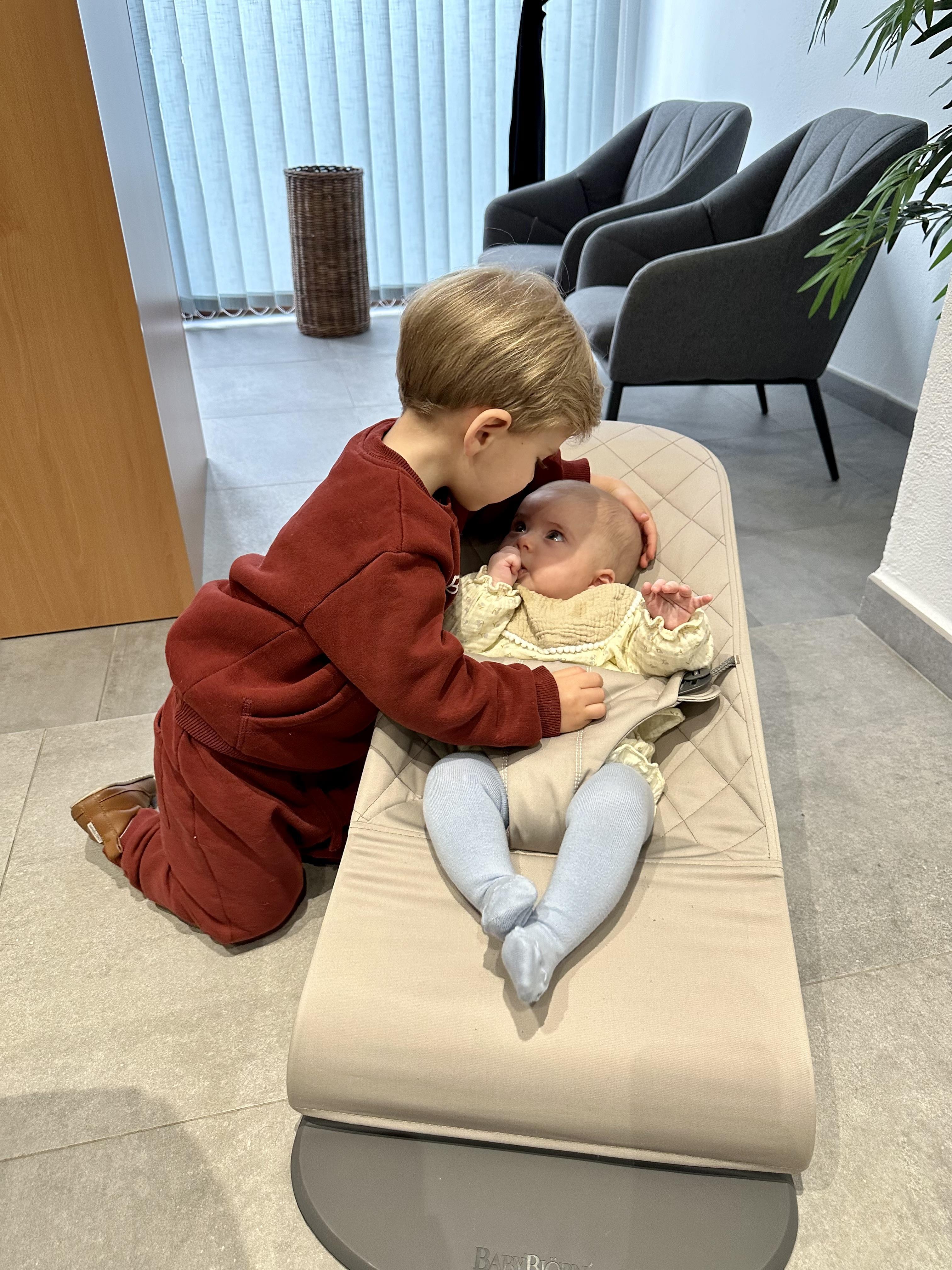A young boy gently touches the head of a baby lying in a beige baby bouncer in a cozy room with modern furniture.