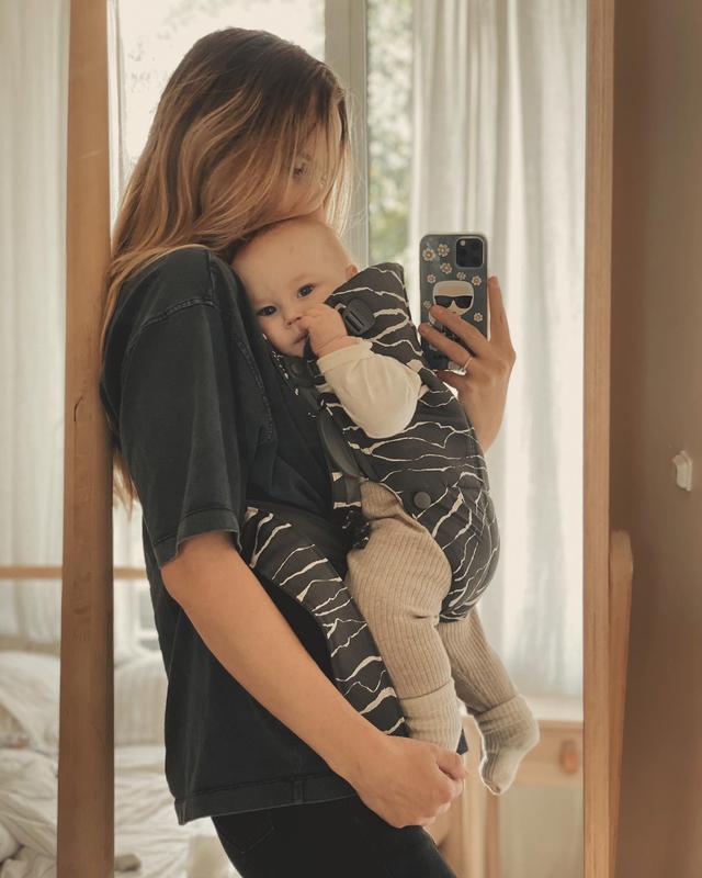 Woman taking a mirror selfie, holding a baby in a patterned carrier. Both face the camera, standing in a softly lit room.