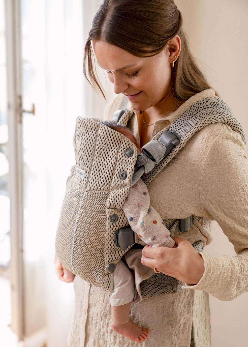 Woman gently holds a baby in a beige mesh carrier, smiling affectionately. The baby wears a colorful polka dot onesie.