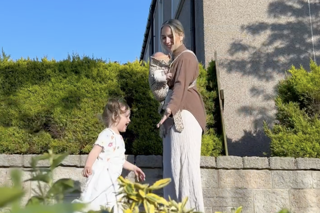 A woman stands outdoors holding a baby, extending her hand to a smiling child in a white dress. The background features a green hedge and a house.
