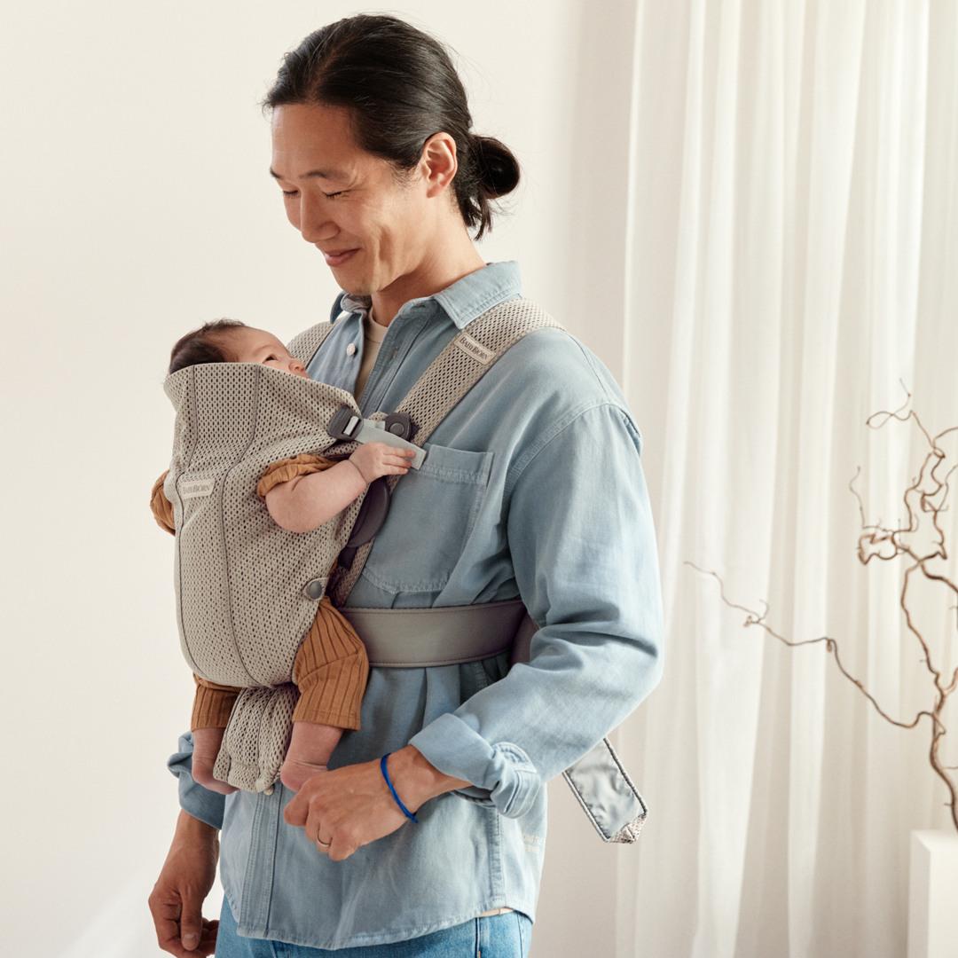 A person in a blue shirt smiles down at a baby in a beige carrier, standing in a softly lit room with white curtains and a decorative branch.