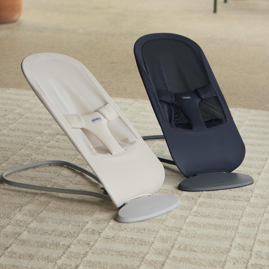 Two baby bouncers on a carpeted floor; one is beige and the other is navy, both with sleek modern designs and harnesses for safety.