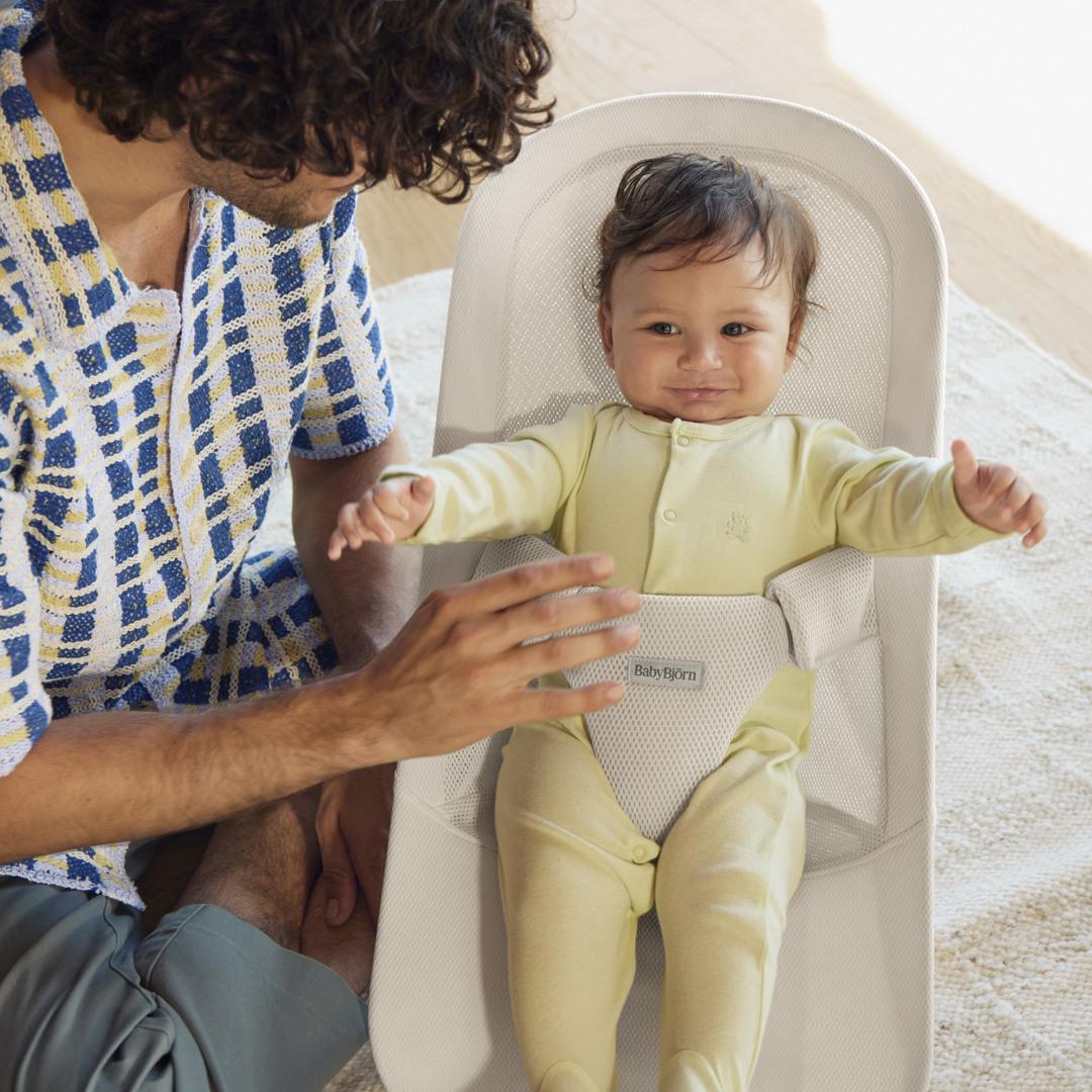 A man with curly hair interacts with a smiling baby in a light yellow onesie lying in a white baby bouncer.