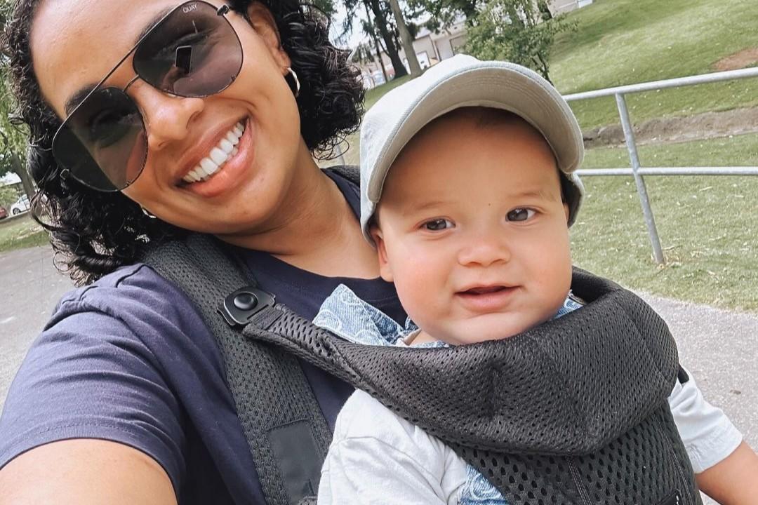 A woman wearing sunglasses smiles while carrying a baby in a front carrier. They are outside in a park with trees and grass in the background.