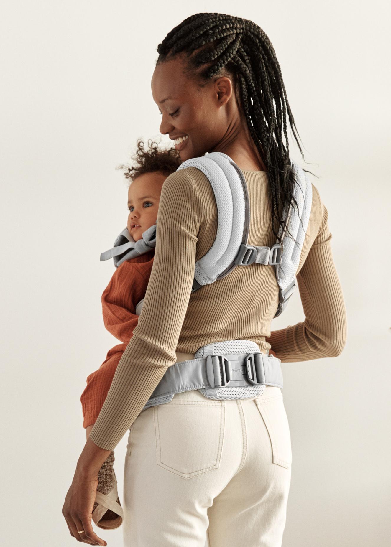 A woman with braided hair holds a baby in an orange outfit using a gray baby carrier.