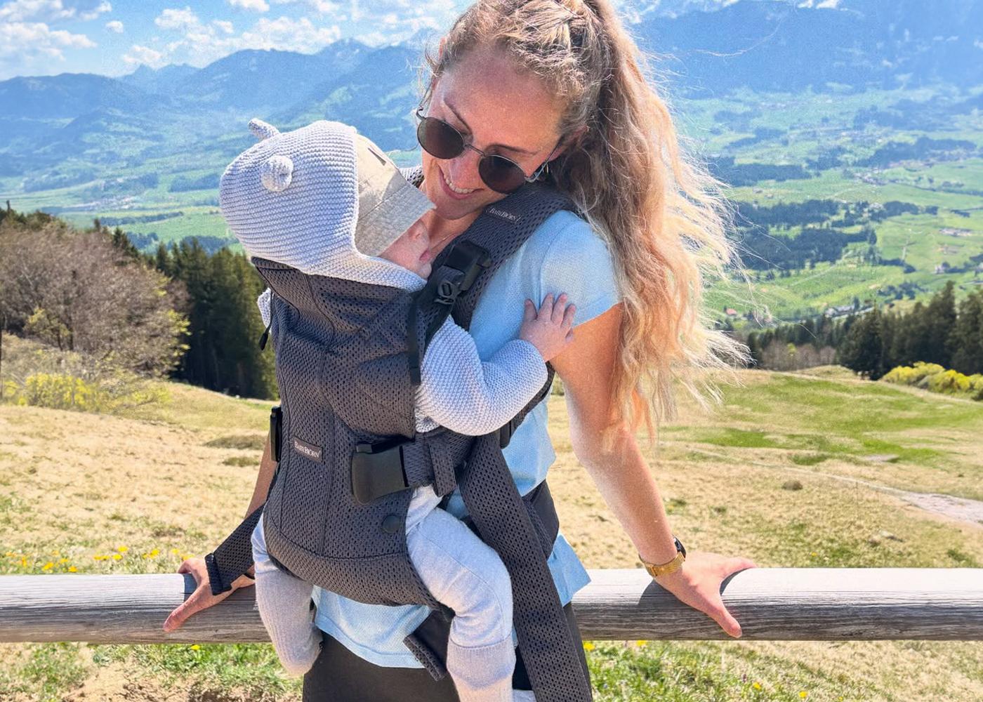 A woman with sunglasses carries a baby in a front carrier, standing by a wooden railing with a scenic mountain view in the background.