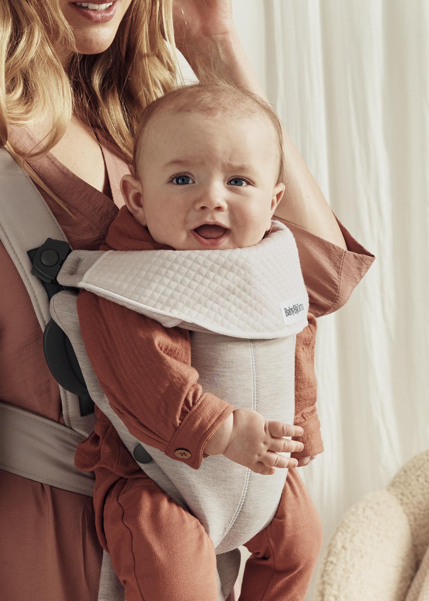 A happy baby in a brown outfit is in a white baby carrier, worn by an adult, smiling at the camera in a softly lit room.