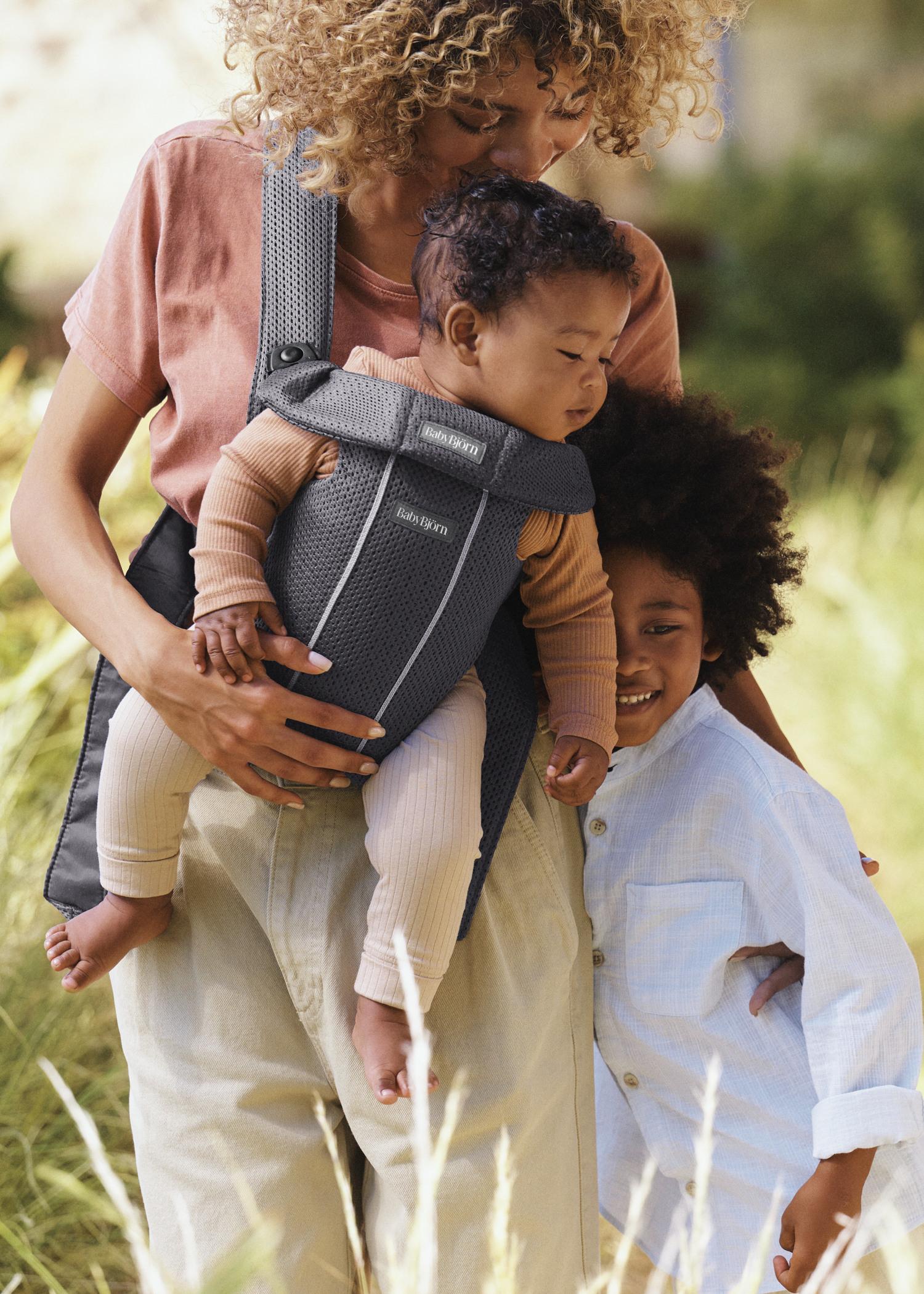 A parent holds a baby in a carrier, with a smiling child hugging them, in a sunny outdoor setting.