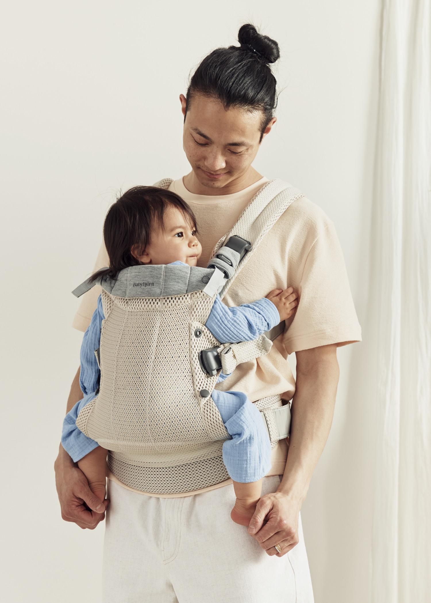 A man in a beige shirt holds a baby wrapped in a light-colored carrier. The baby wears a blue outfit and looks to the side.