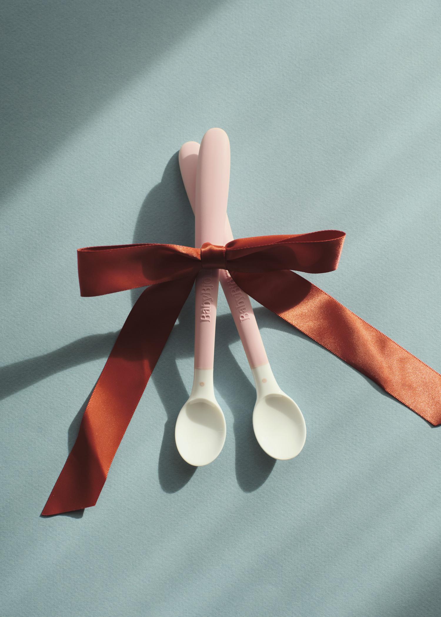Two pink baby spoons tied together with a red ribbon on a light blue background, casting soft shadows.