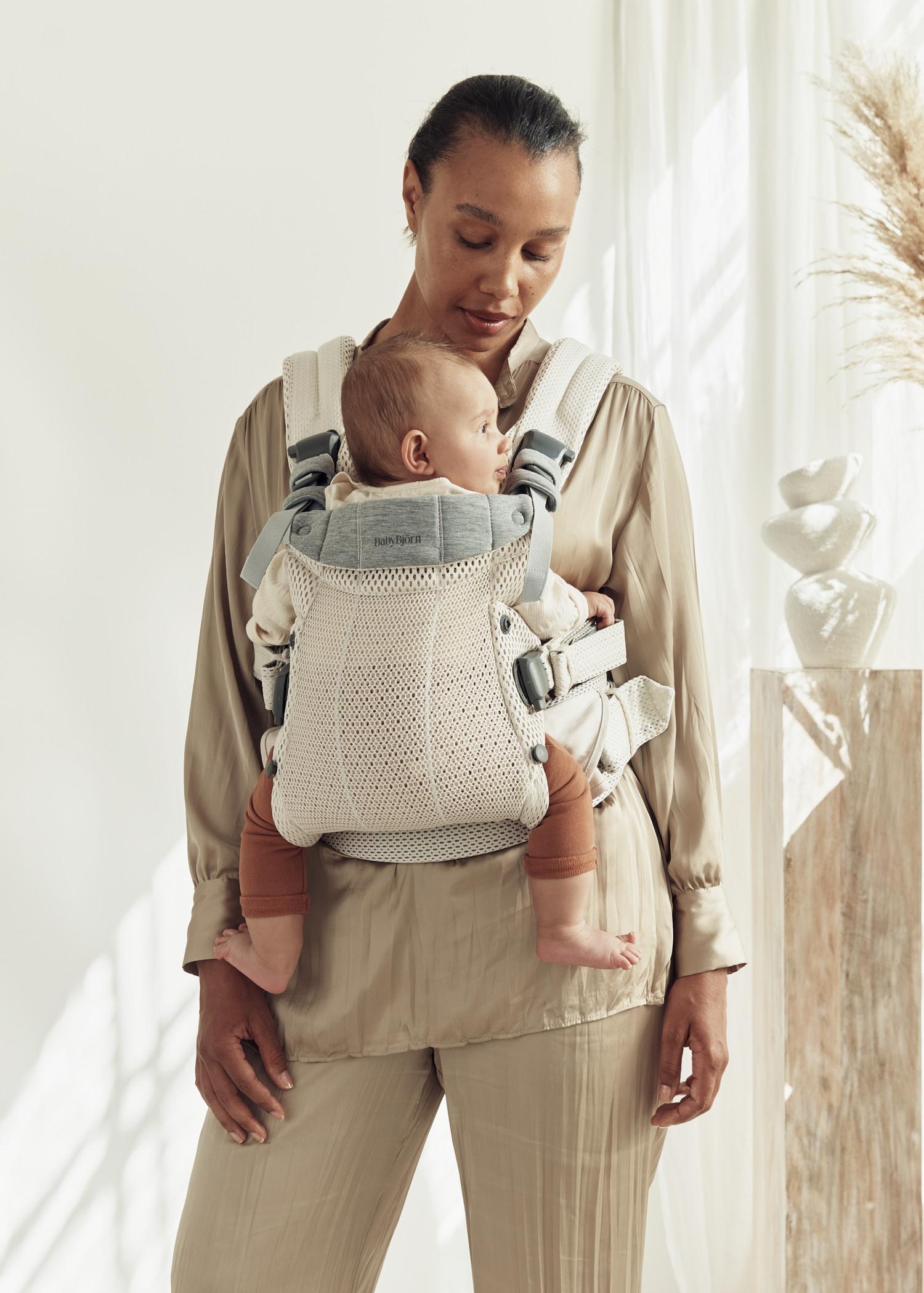 A woman in a beige outfit carries a baby in a mesh baby carrier. The room is softly lit with neutral decor and pampas grass in the background.