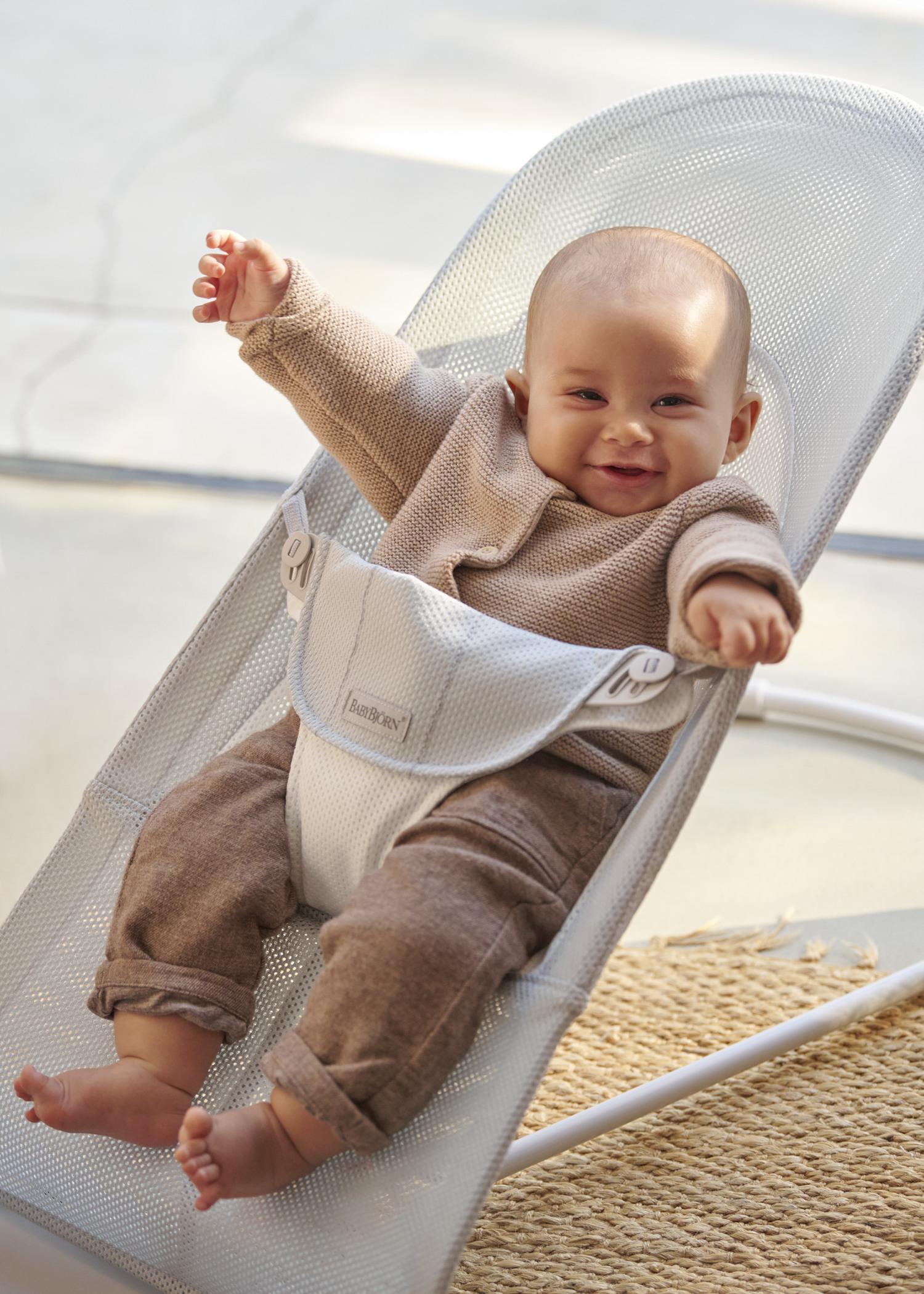 Smiling baby in a beige outfit sitting in a white baby bouncer, arms raised, on a light-colored floor with a woven rug nearby.
