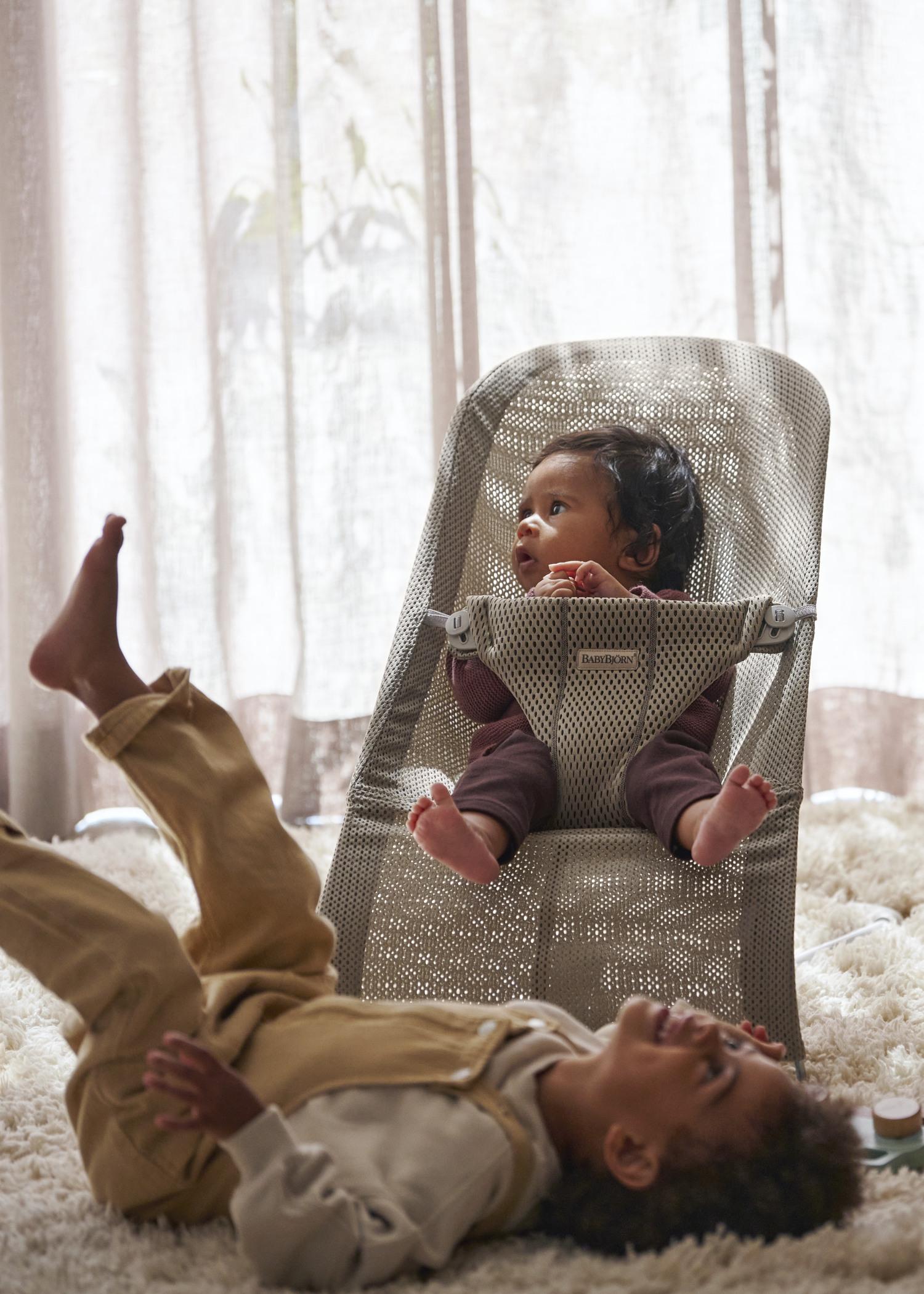 Two young children in a cozy room; one sits in a baby bouncer, while the other lies on a fluffy rug, both appearing playful and relaxed.