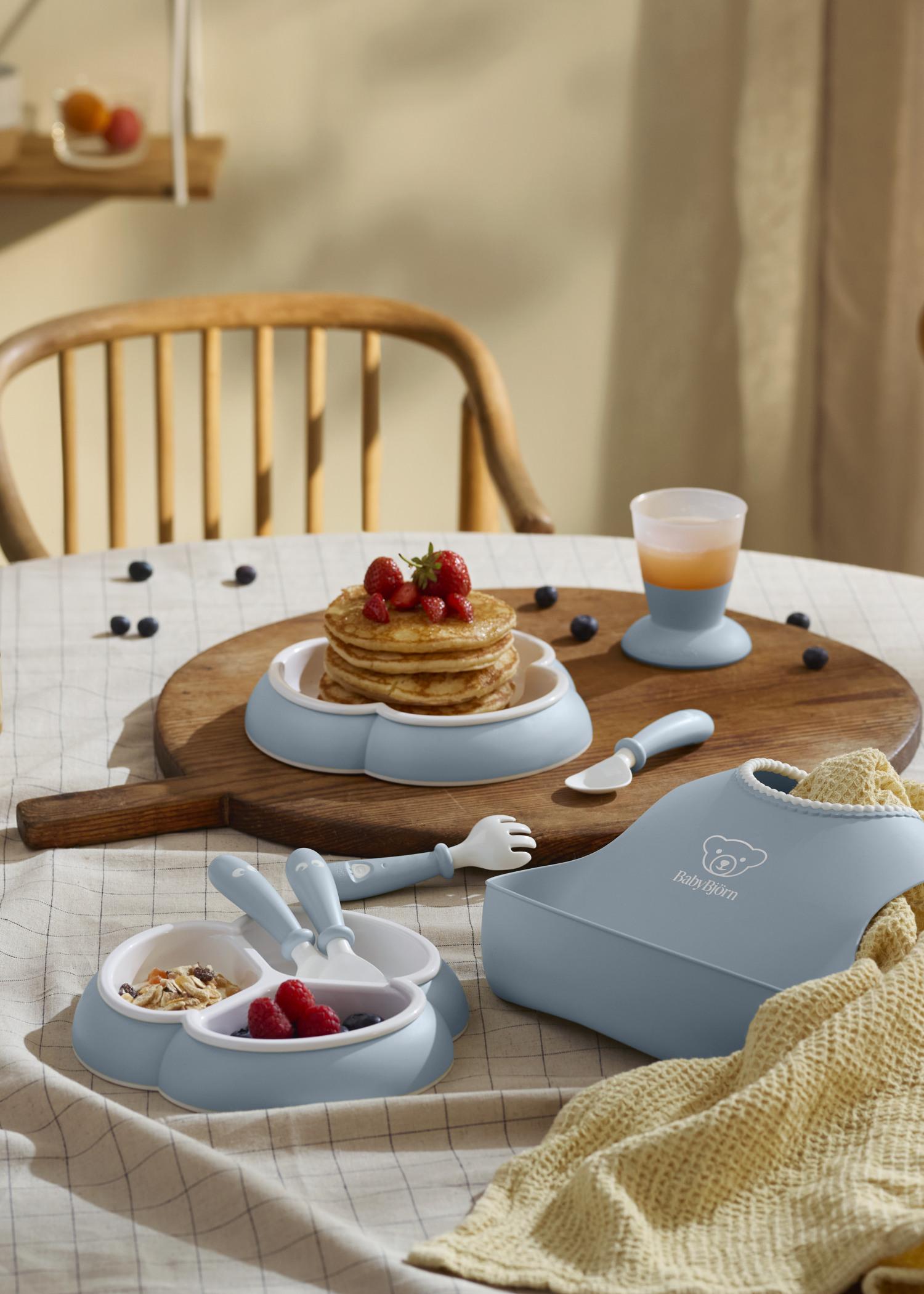 A cozy breakfast setup with pancakes topped with strawberries, a baby bib, utensils, and a cup on a table with a checkered cloth.