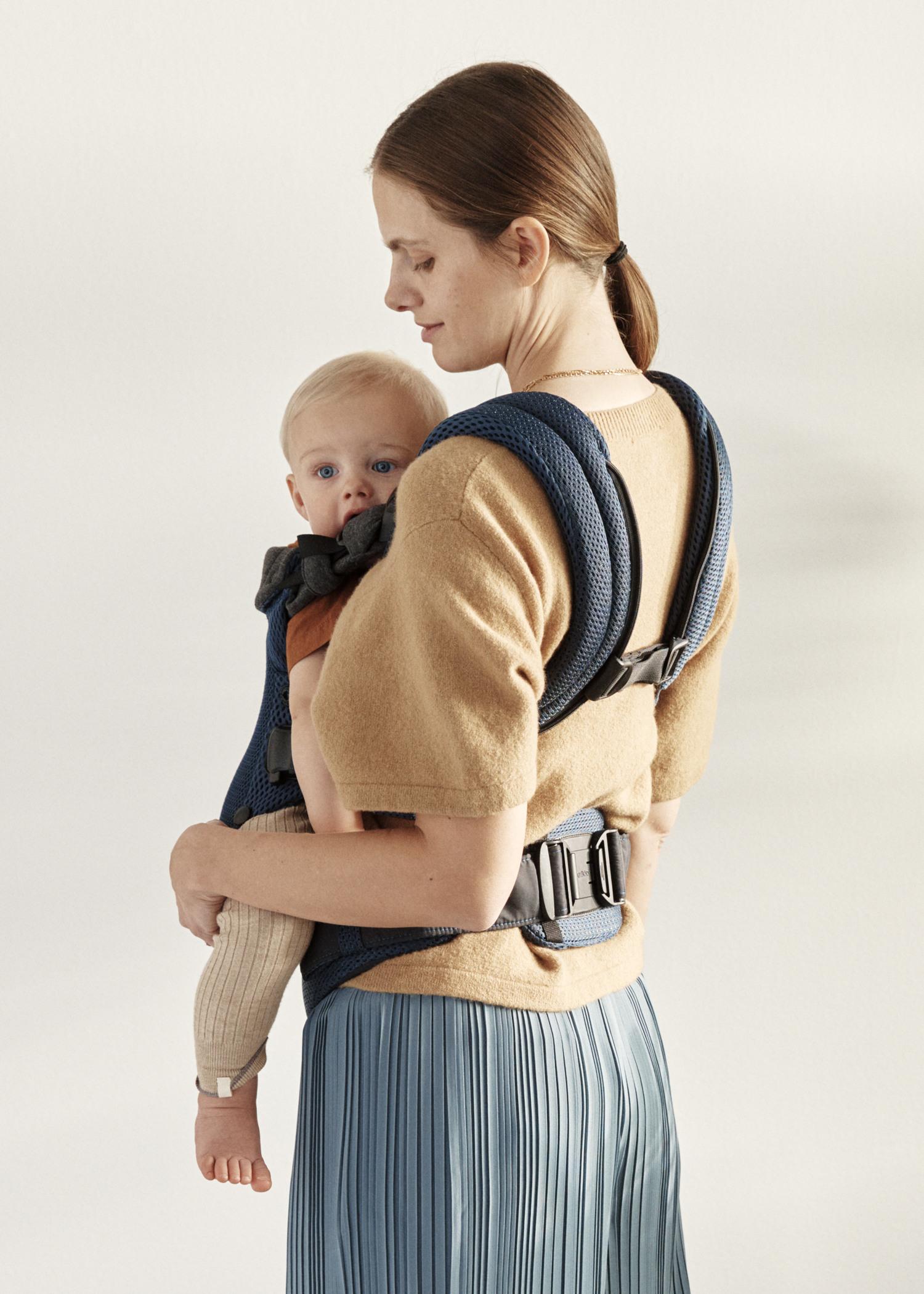 Woman with brown hair wearing a beige top and blue skirt, carrying a baby in a navy blue carrier against a neutral background.